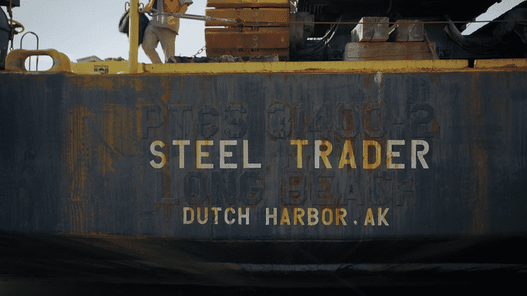 Close-up of a rusty steel cargo ship's hull with the words "Steel Trader" and "Dutch Harbor, AK" painted on it, indicating it is docked at Dutch Harbor, Alaska.
