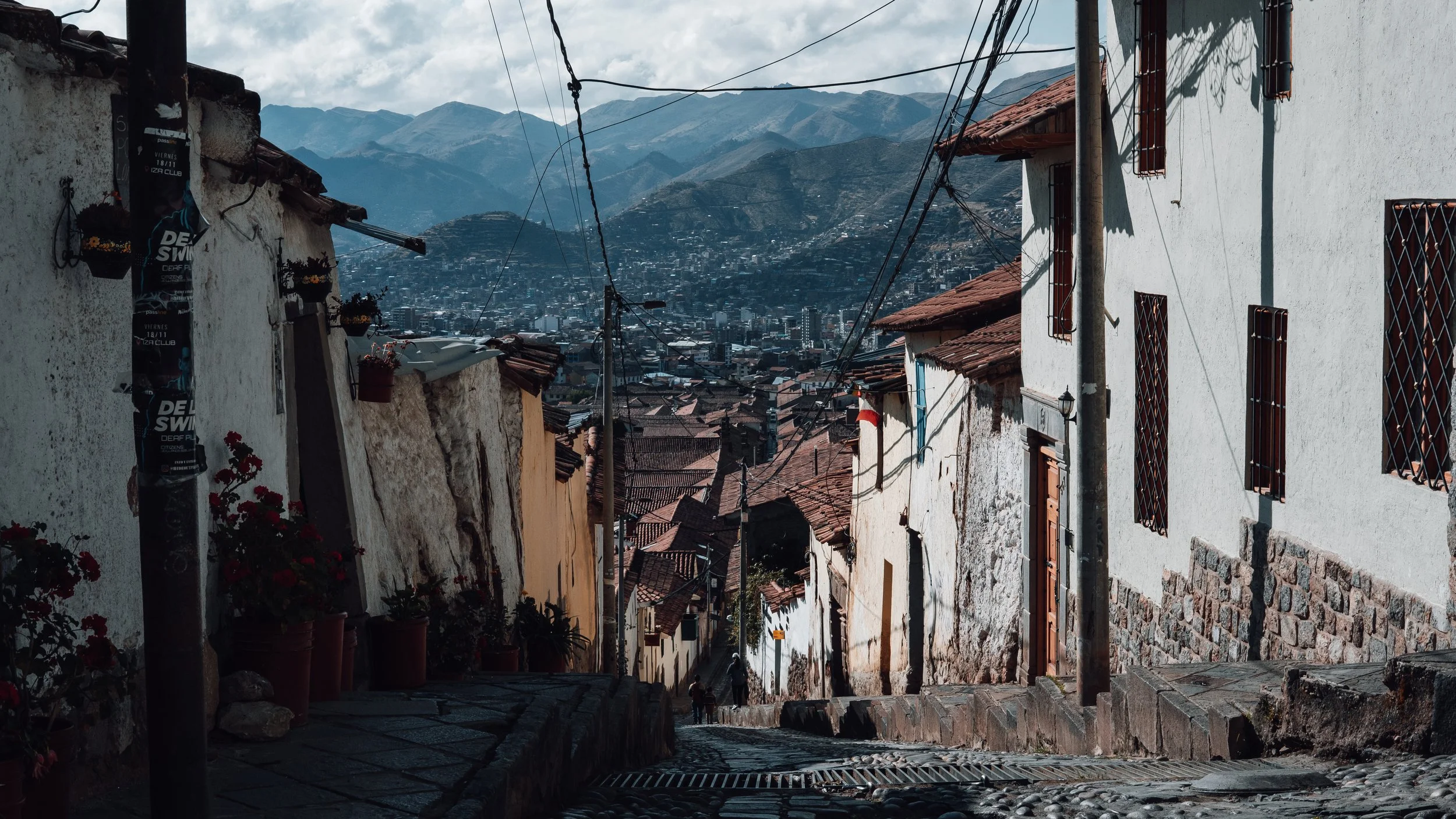 A narrow, cobblestone street slopes downward in a hillside town with white and beige buildings on either side. Wires crisscross above, and in the background, there are mountain ranges with a cityscape at the base.