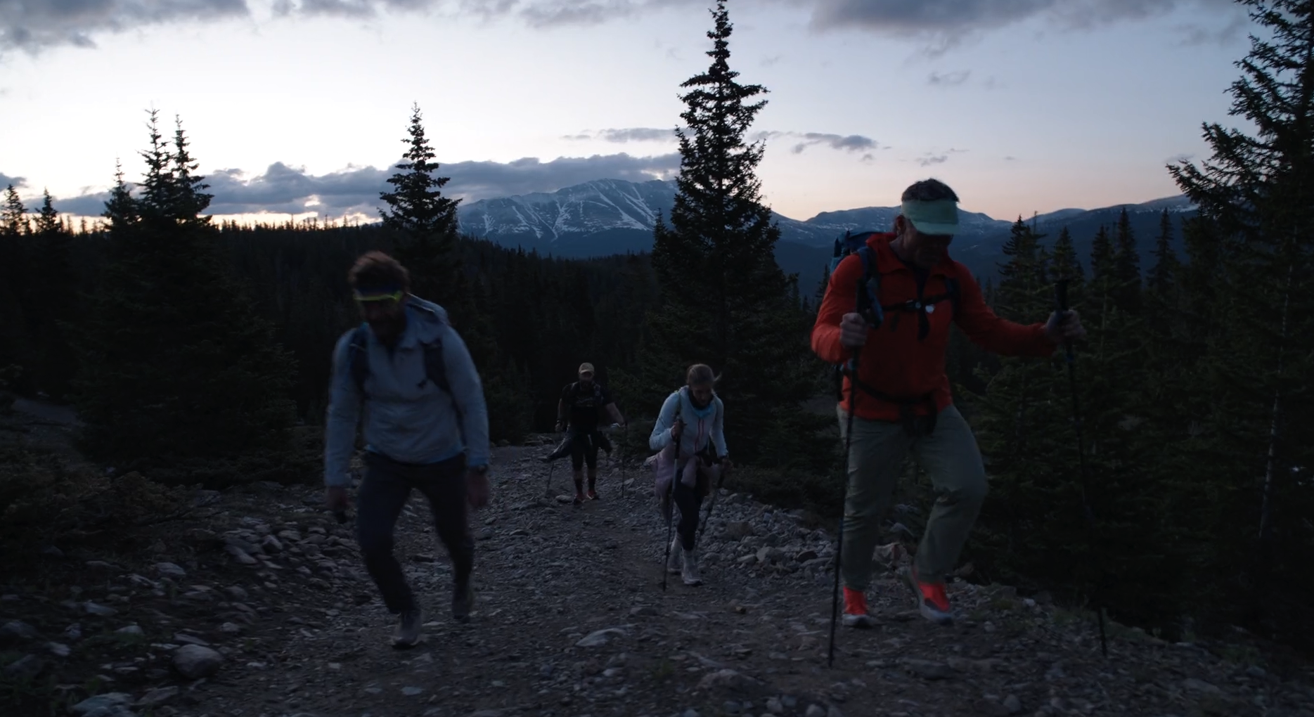 Four hikers walking on a rocky trail in a forested mountain landscape during dusk or dawn, with snow-capped mountains in the background.