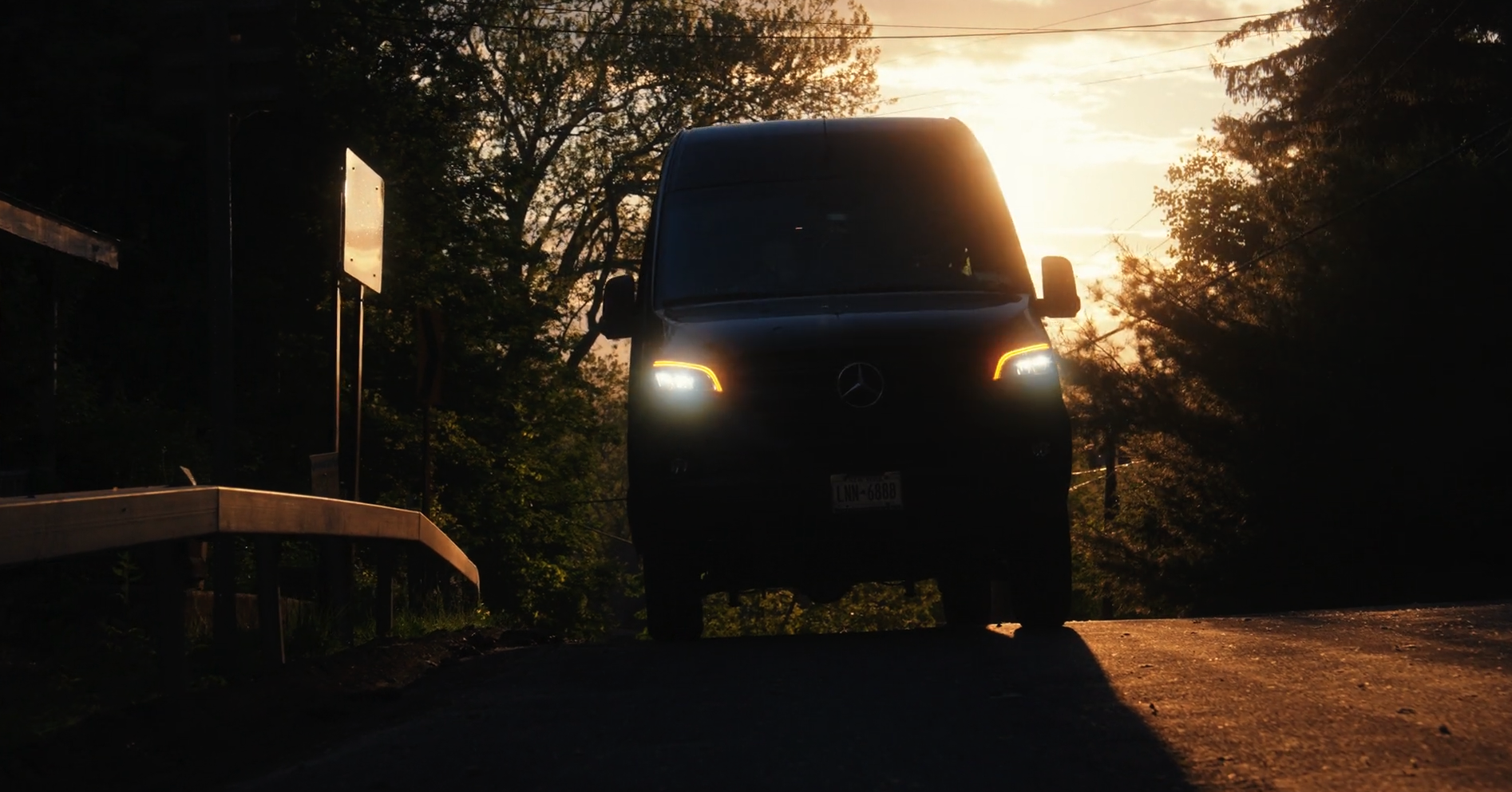 Black Mercedes van with headlights on, parked on a road during sunset with trees and sky in the background.