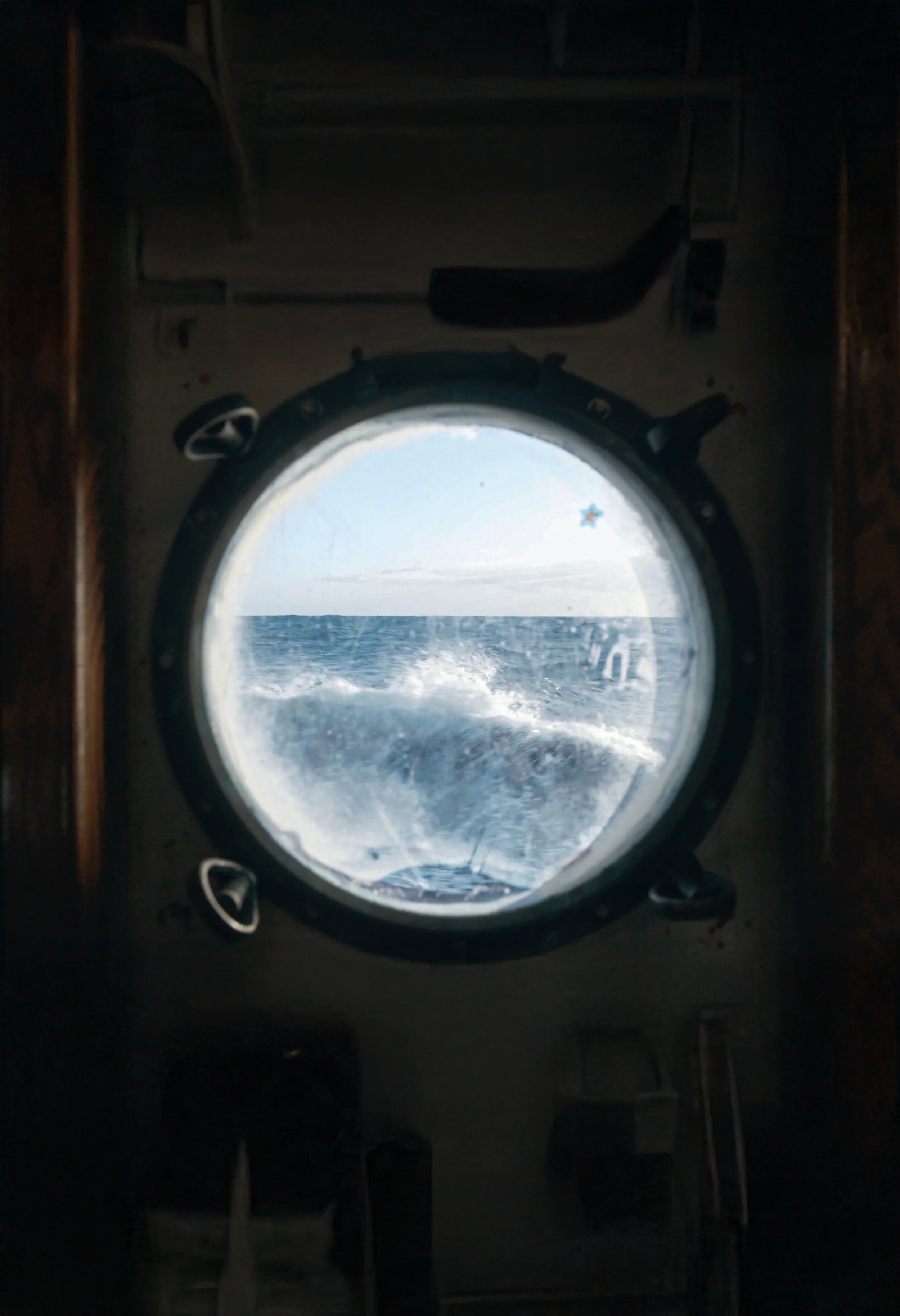 View of the ocean through a ship's porthole, with waves and a boat wake visible.