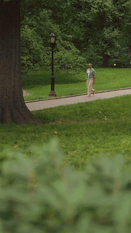 A man walking on a sidewalk in a park with green grass, trees, a lamp post, and a yellow ball in the background.