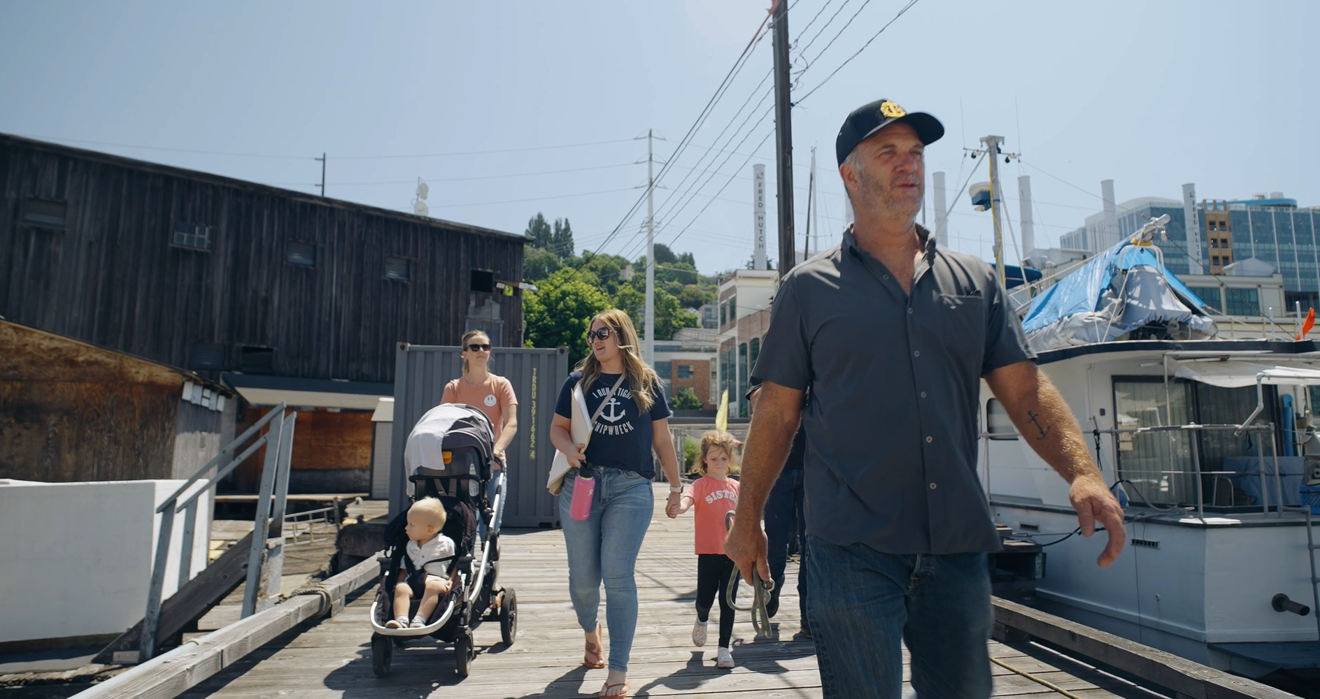 People walking along a marina pier on a sunny day, with boats docked on the right and a dark wooden building on the left.