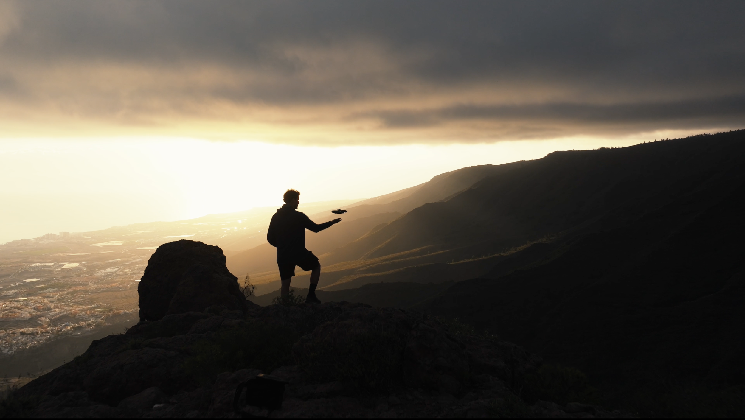 Silhouette of a person standing on a rocky hill during sunset, holding a drone in their hand, with mountains and a valley in the background under dark clouds.
