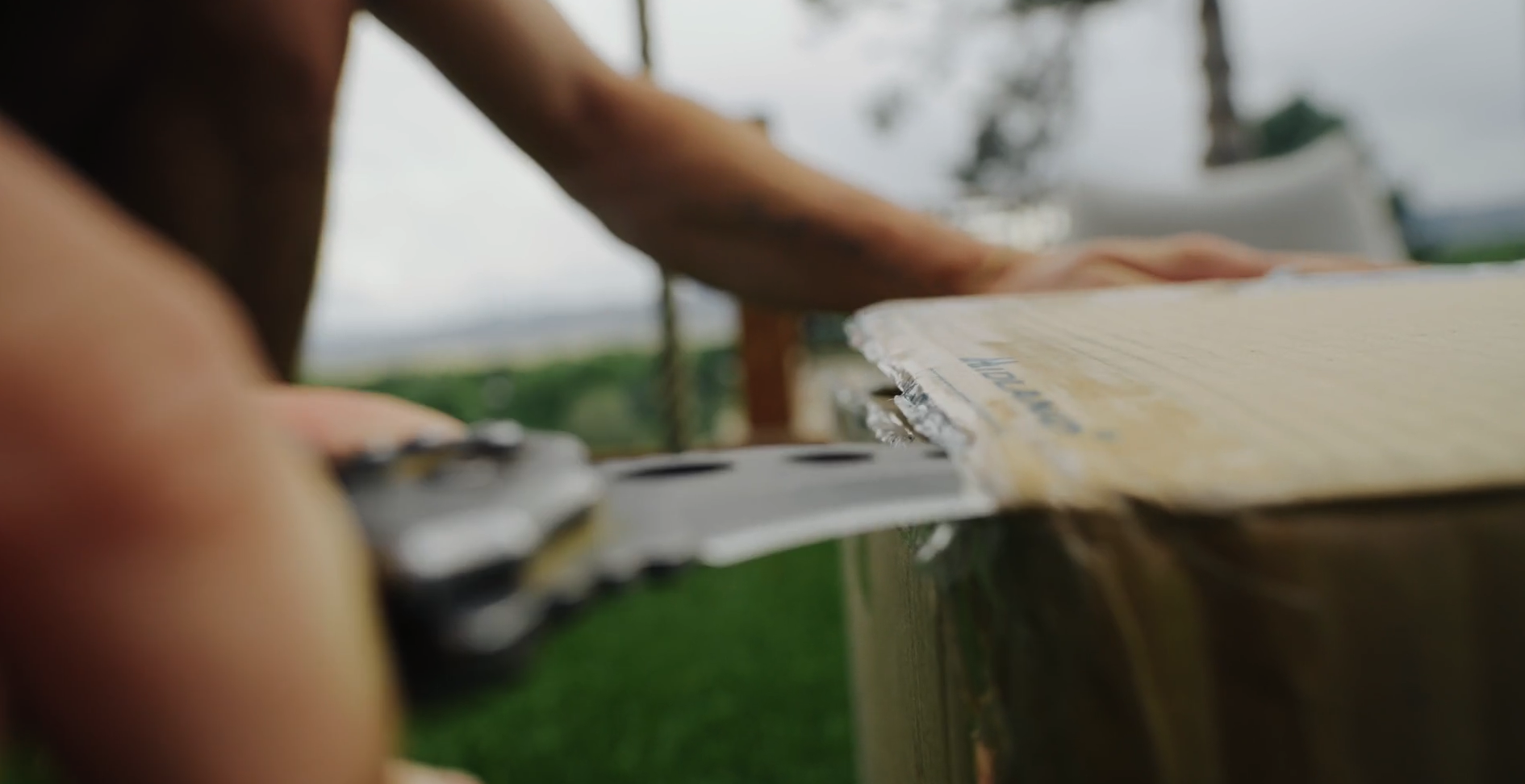 Close-up of a person's hand cutting a cardboard box with a handsaw outdoors, with a blurred green landscape and trees in the background.
