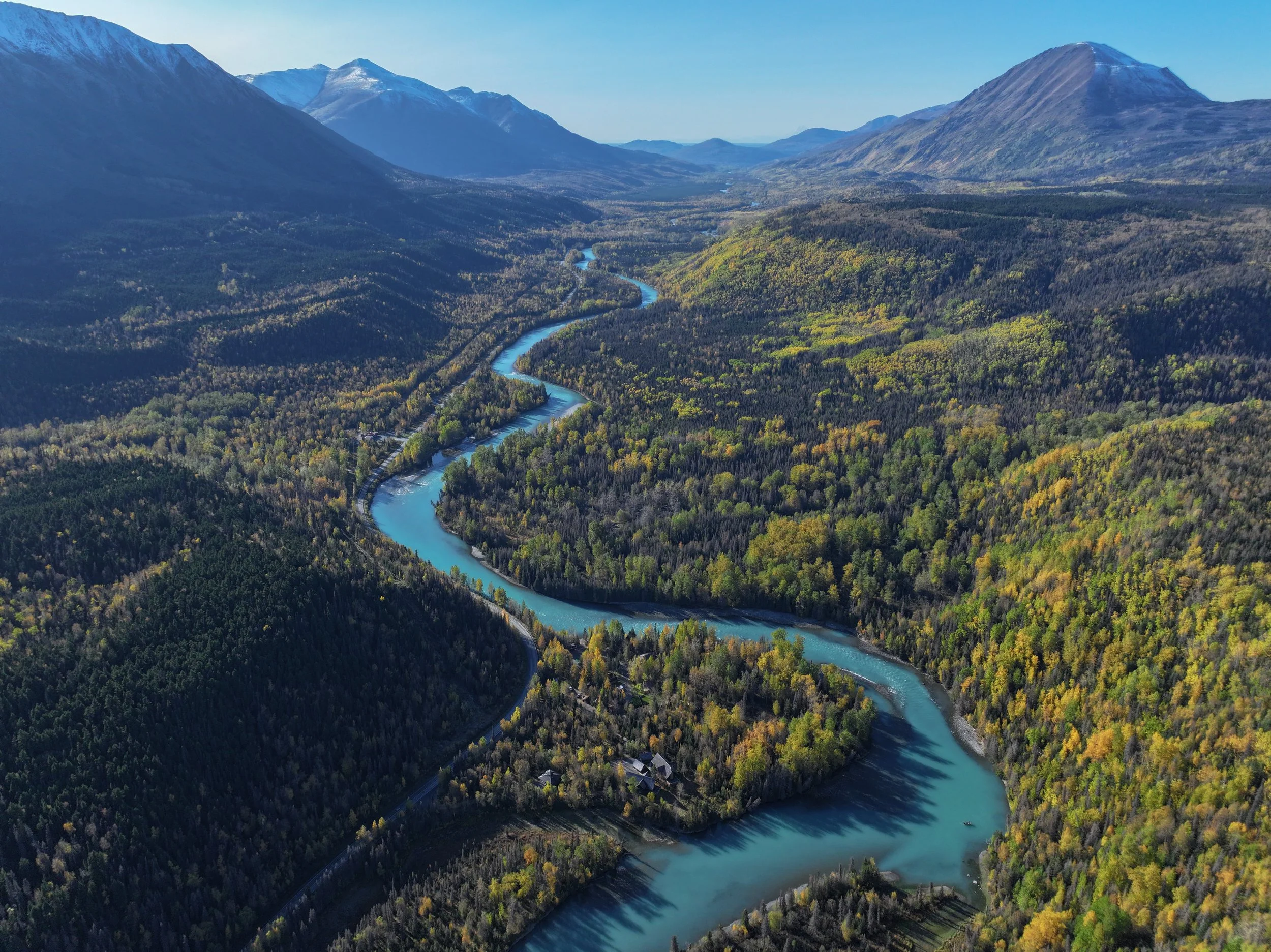Aerial view of a winding river flowing through a lush forested valley surrounded by mountains with some snow-capped peaks in the distance during daytime.