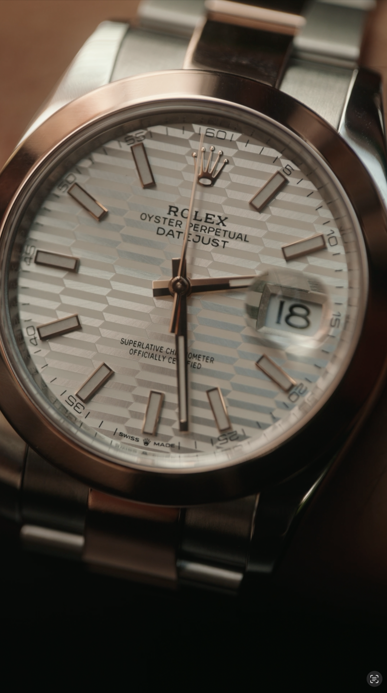 A close-up of a Rolex Oyster Perpetual Datejust wristwatch with a white textured dial, rose gold hour markers, and a date window showing the 18th.
