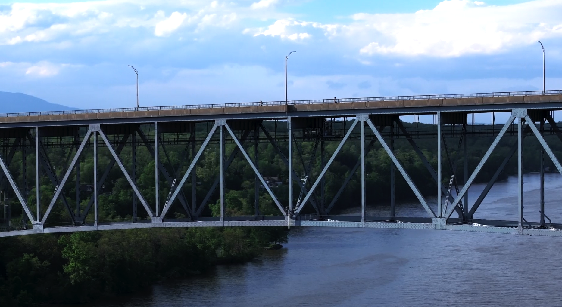 A scenic view of a steel truss bridge spanning over a river, with lush green trees on the riverbanks and a cloudy sky above.