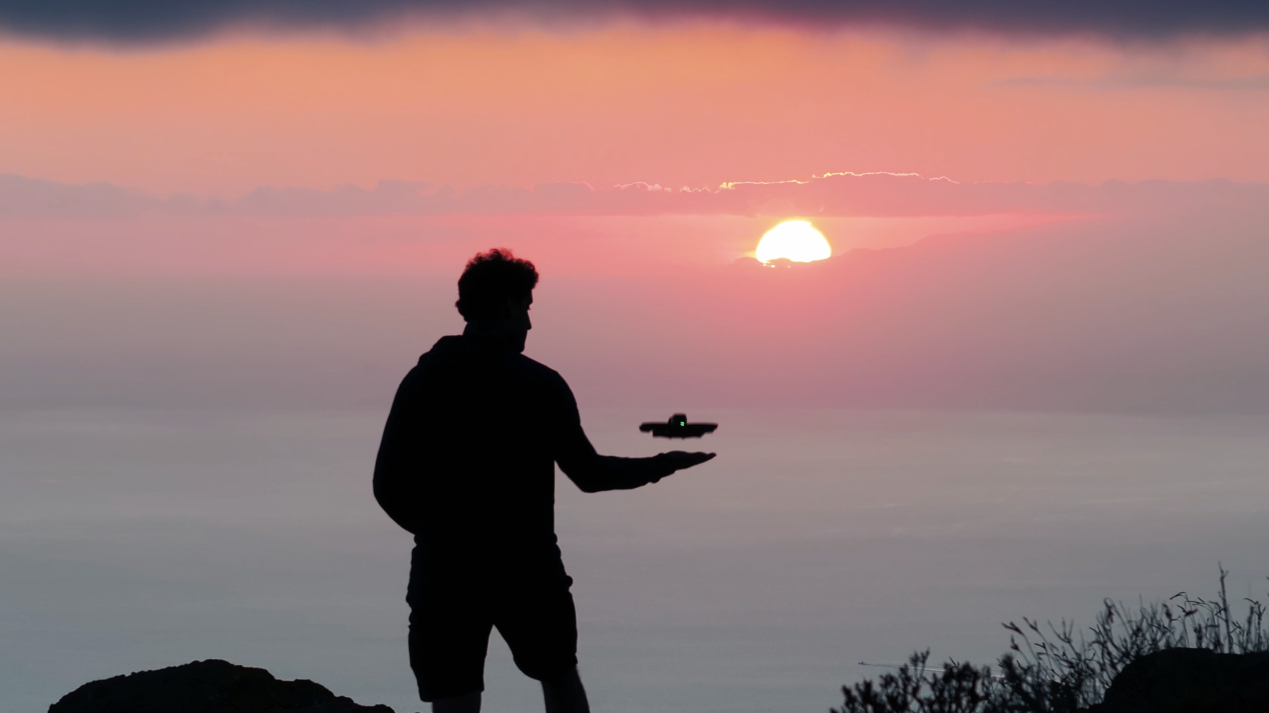 A silhouette of a person holding a drone near the ocean at sunset, with the sun setting behind clouds in pink and orange hues.