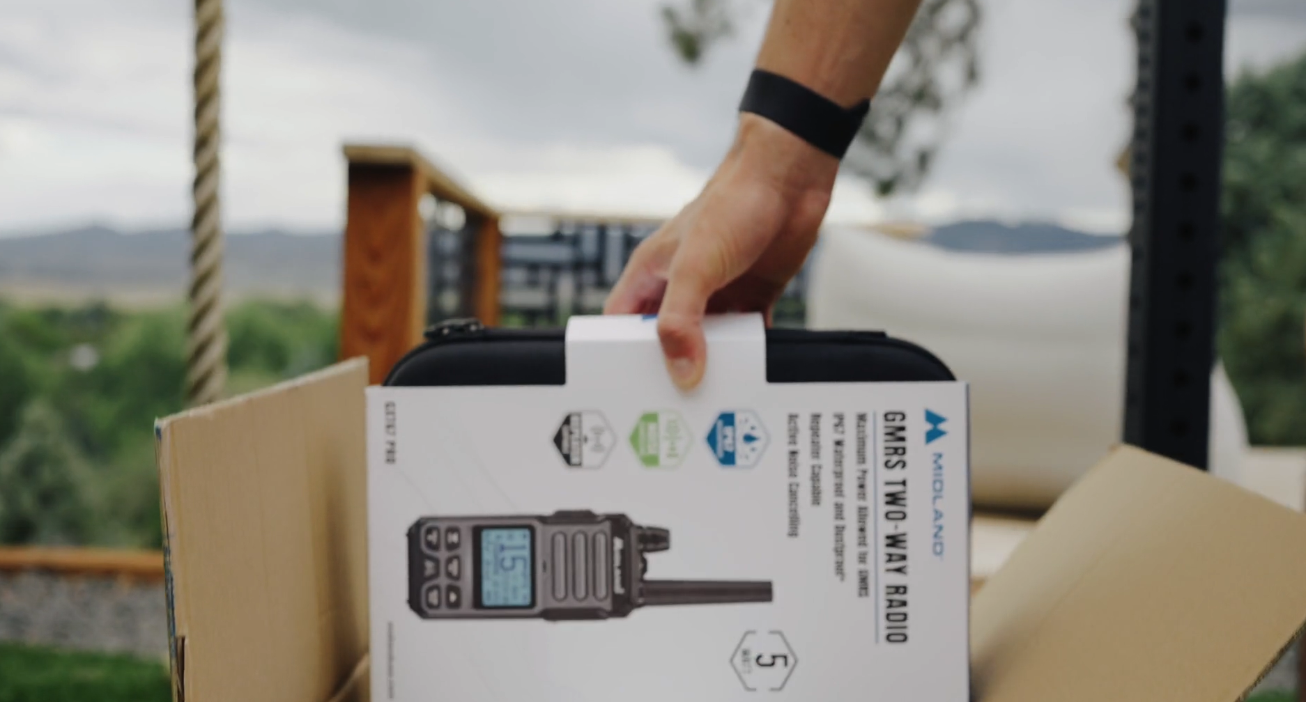 Hand holding a Midland Walkie-Talkie Radio in front of a cardboard box outdoors with trees and a cloudy sky in the background.