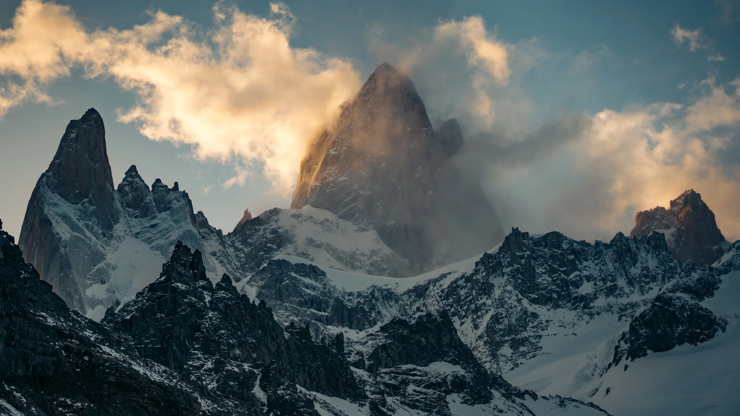 Snow-covered jagged mountain peaks under a cloudy sky with sunlight illuminating the clouds.