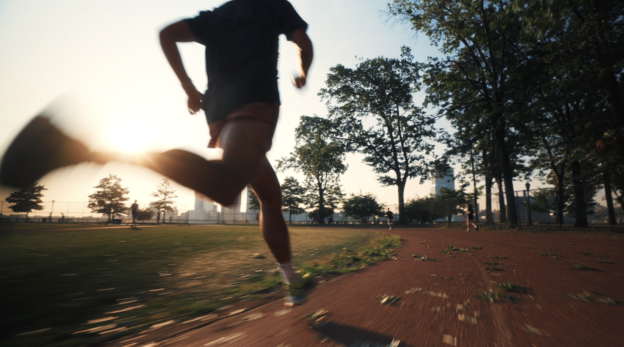 Person running on a track in a park during sunset with trees and city buildings in the background.
