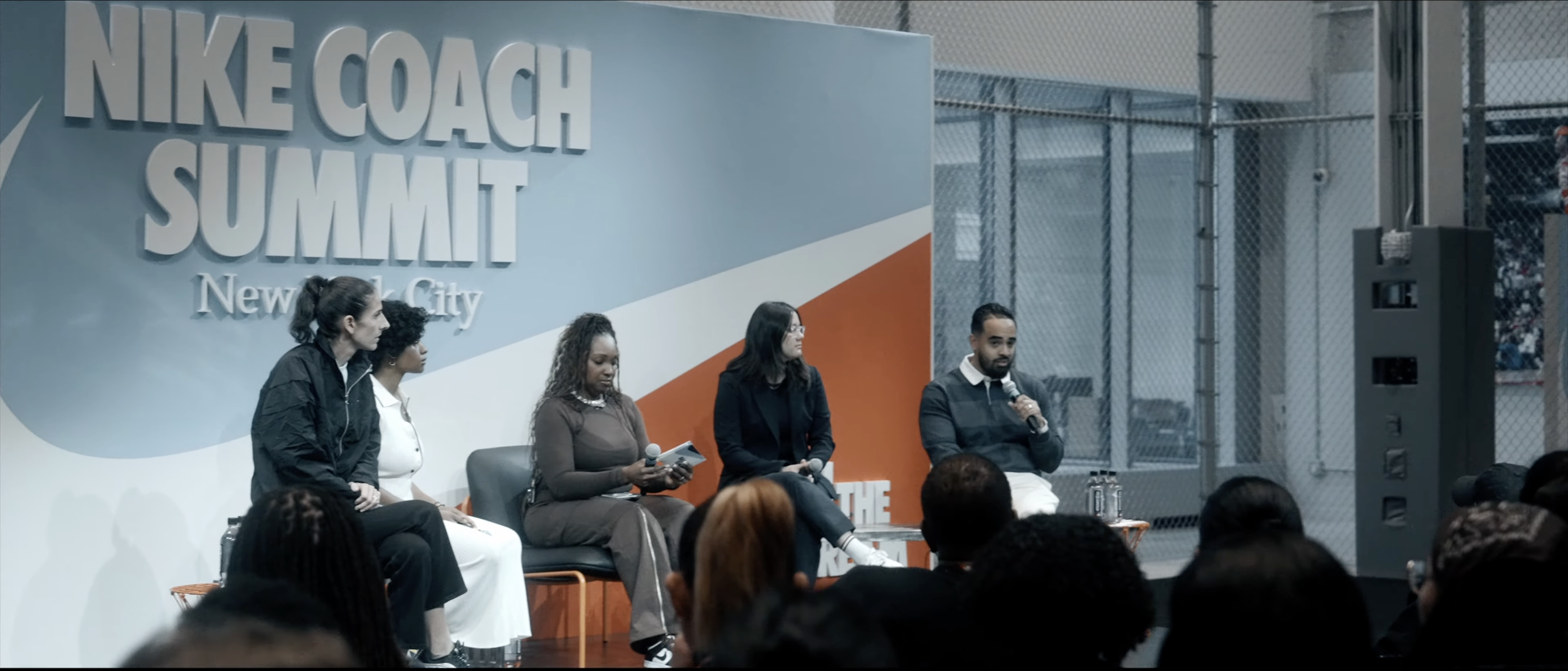 Panel of five diverse individuals speaking at the Nike Coach Summit in New York City, seated on stage in front of a large Nike Coach Summit banner, with an audience watching.