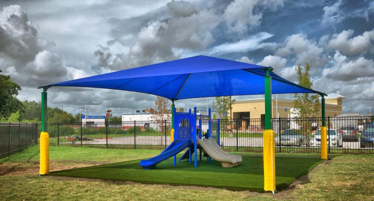 A small outdoor playground with a blue shade sail overhead, featuring two slides, one blue and one beige, on a grassy area with a black metal fence in the background.