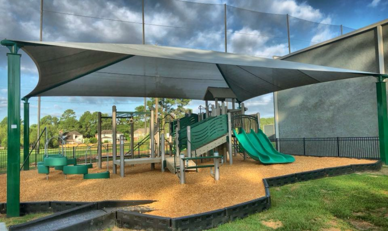 Children's playground with a shade sail, slides, and climbing structures on a rubberized surface, surrounded by a black safety fence.