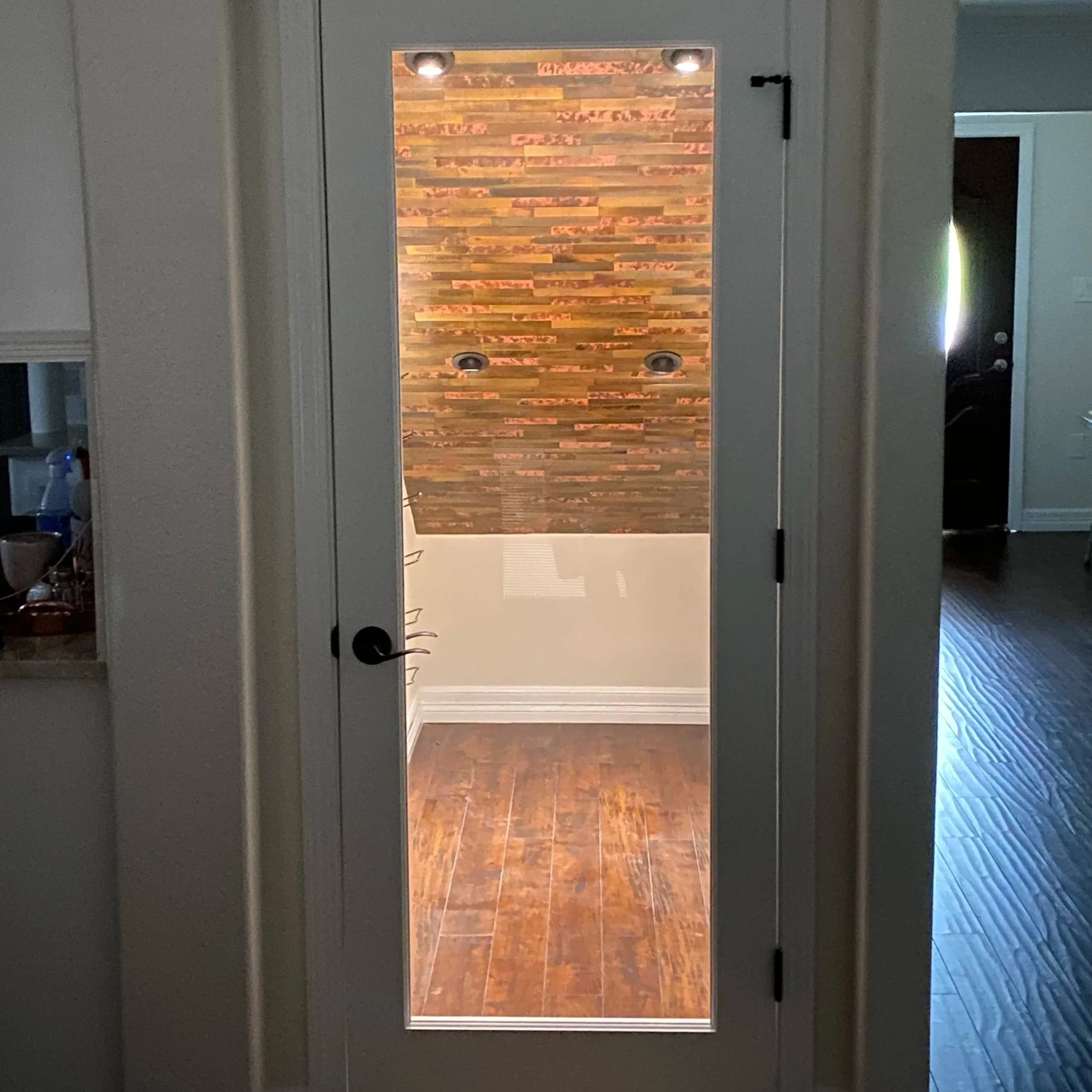 A mirror on a door reflects a room with a wooden floor and a wood-paneled accent wall, with recessed lighting.
