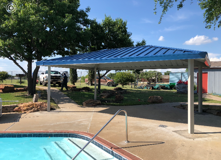 A covered poolside shade structure with blue metal roof, supported by white columns, located next to a swimming pool with a metal handrail, in a backyard with trees and green lawn, and some parked cars in the background.