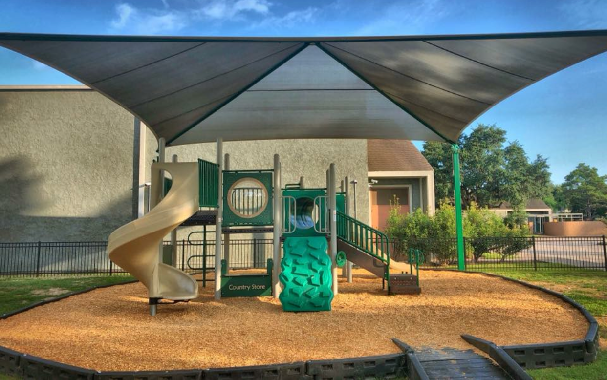 Children's playground with a slide, climbing wall, and bridge, covered by a large shade sail, located in a neighborhood park.