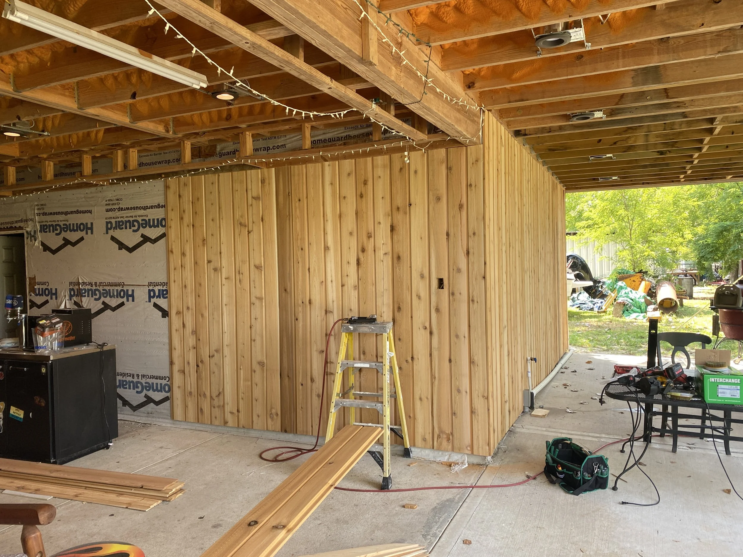 Construction site with unfinished wooden room and tools, partially open to outside with trees and greenery.