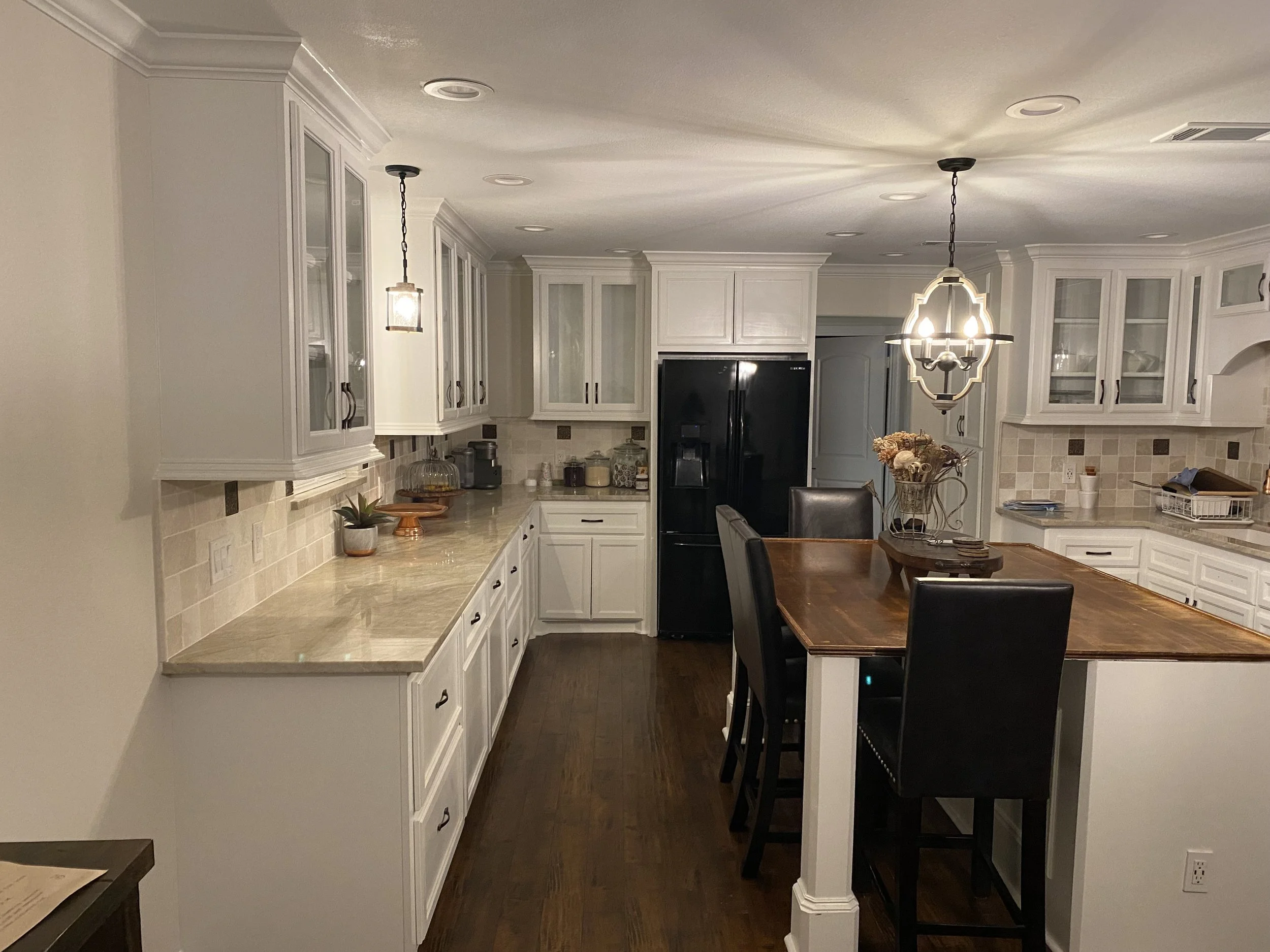 A spacious kitchen with white cabinets, a wooden dining table with black chairs, and a black refrigerator. There are two hanging light fixtures and a decorative arrangement on the table. The countertops are beige with various small kitchen items.