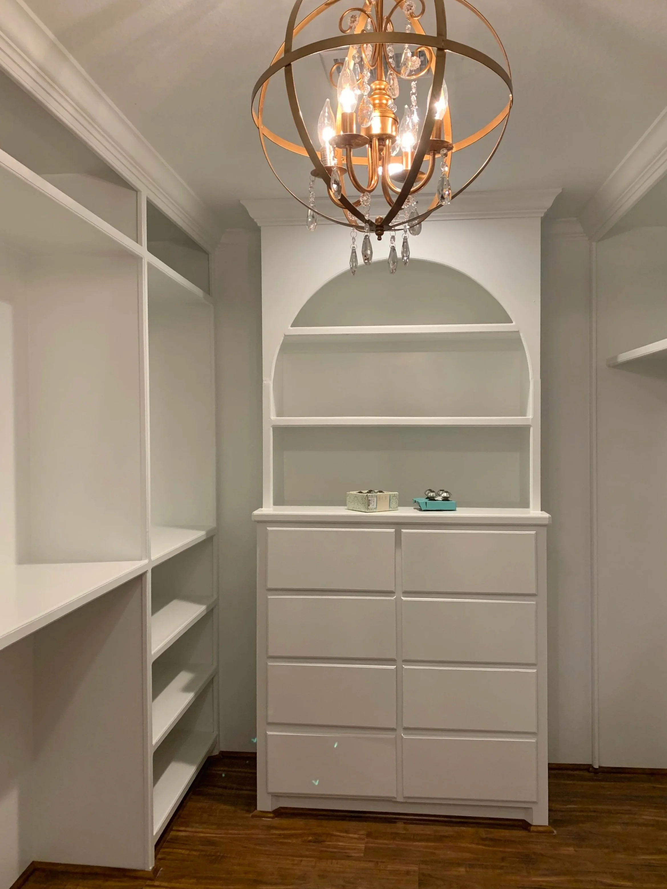 Empty built-in white shelves and drawers in a closet with a chandelier hanging from the ceiling.