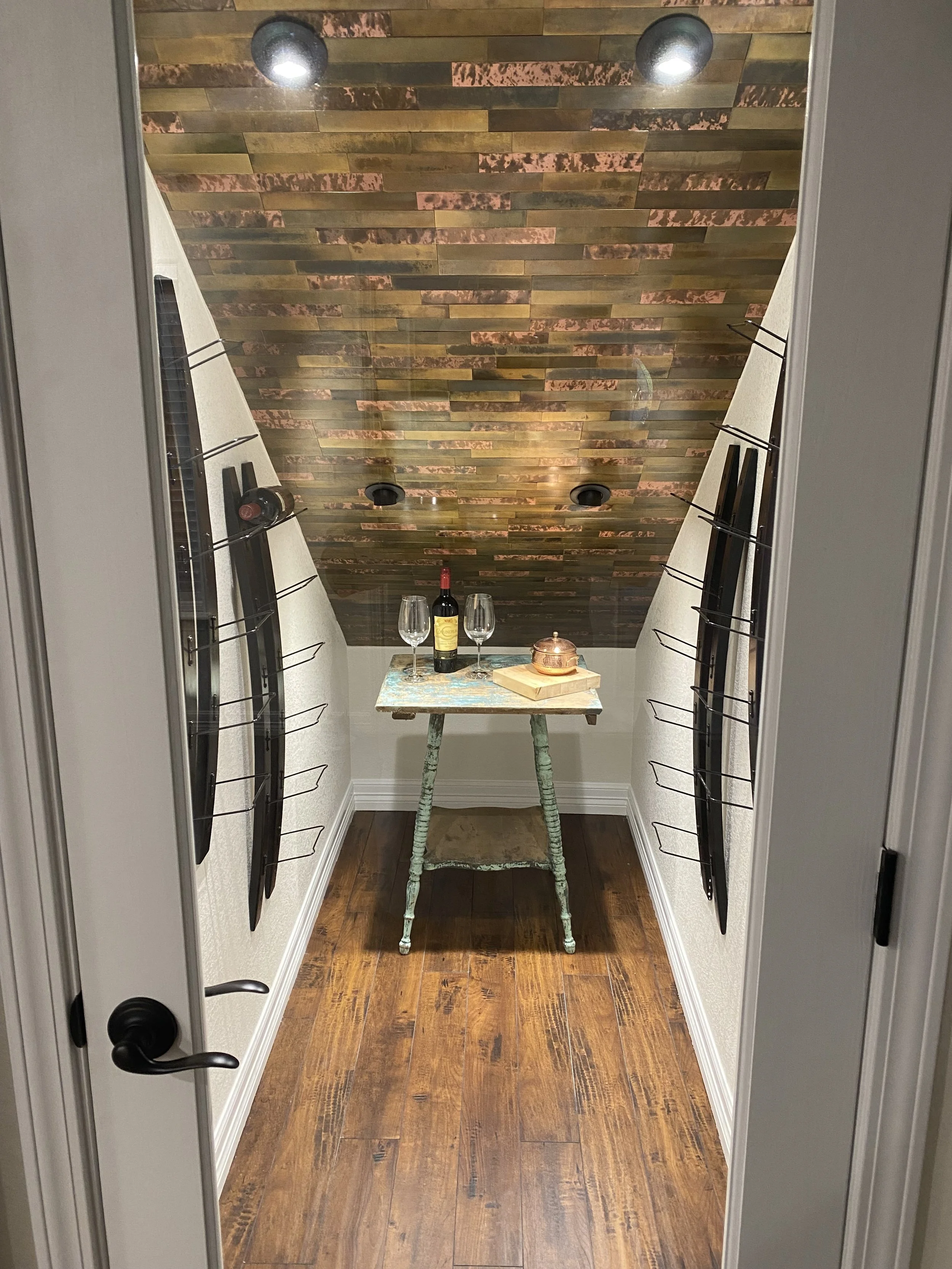 A small wine cellar with a wooden ceiling, white walls, and a wooden floor. A rustic table in the center with a bottle of wine, two wine glasses, and a copper pot. Black metal racks on the walls hold wine bottles.