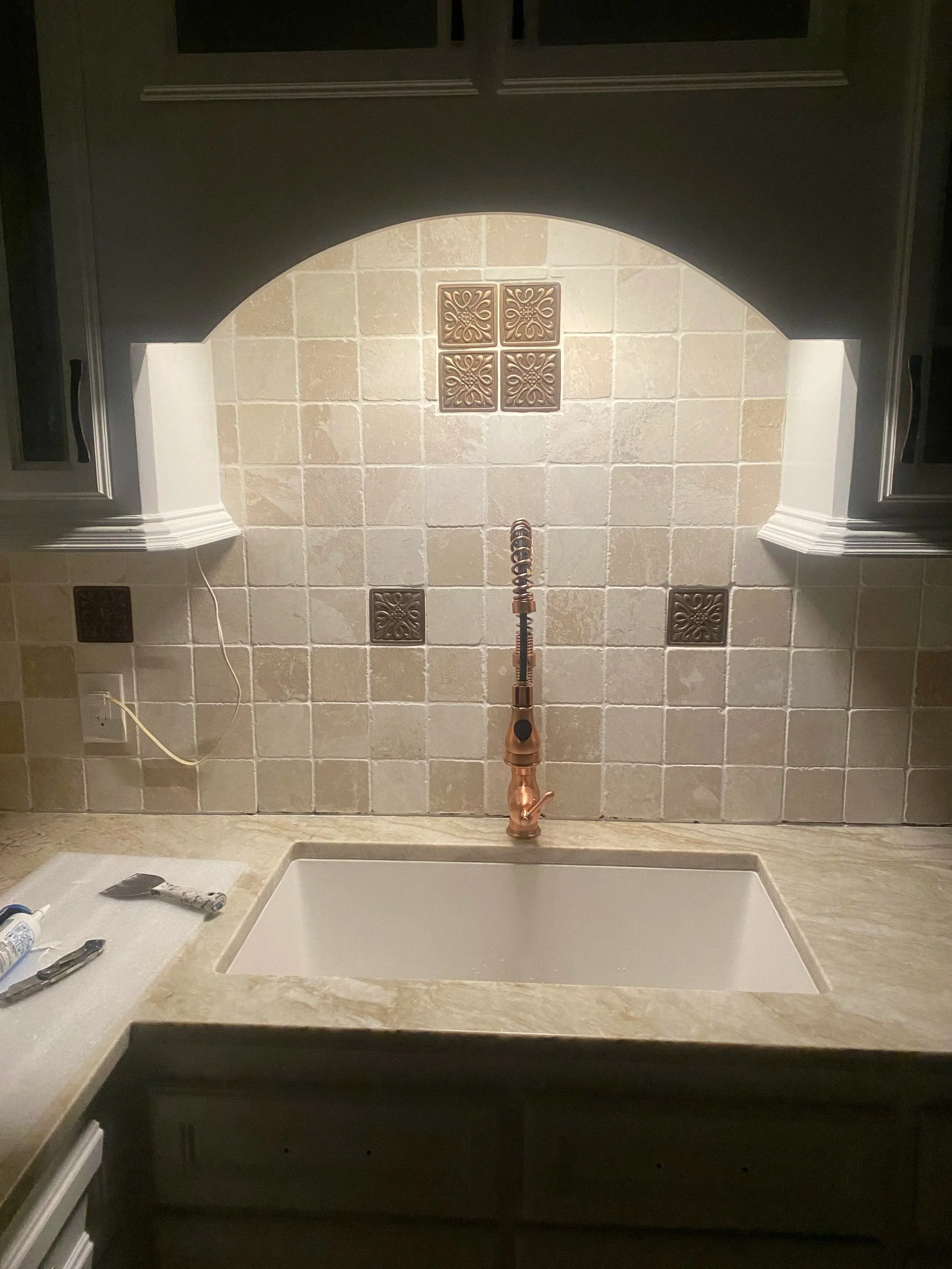 Kitchen sink with a copper faucet in a tiled backsplash area, surrounded by dark green cabinets and a beige countertop.