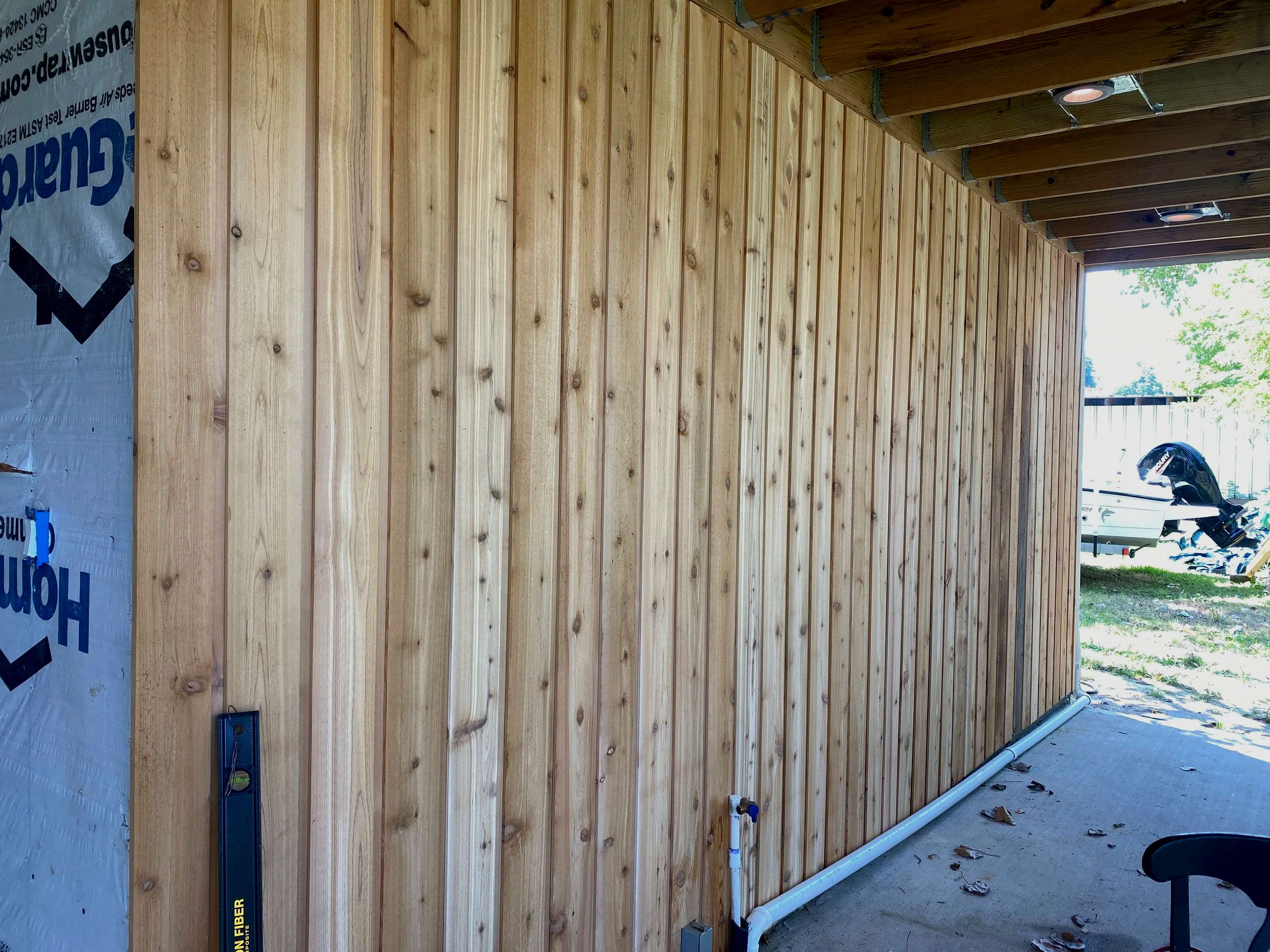 Wooden wall under construction in a building, with a level tool hanging on the left side and exterior view with boats and trees visible through an open side.