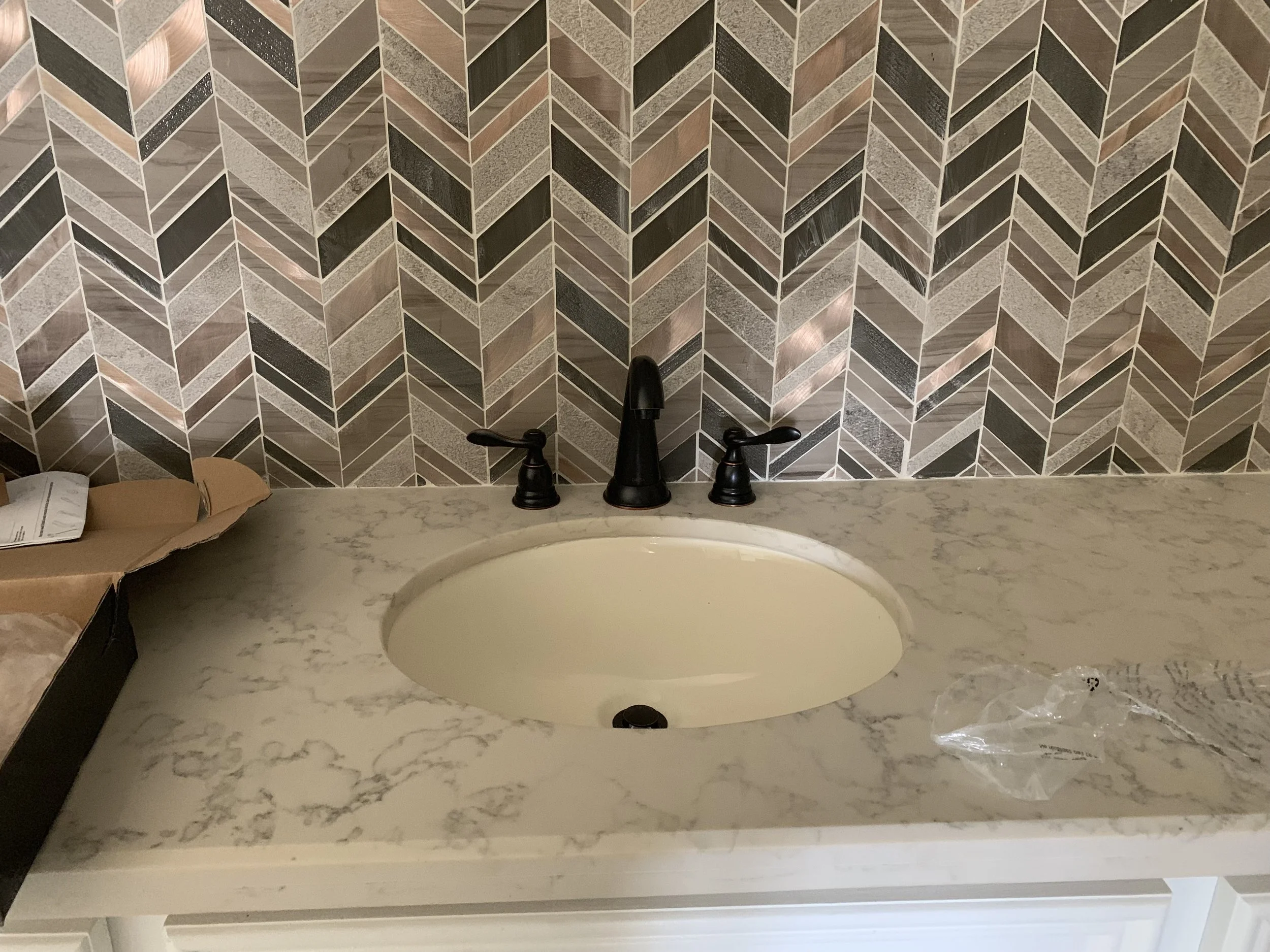 Bathroom sink with a marble countertop, black faucet, and a geometric tile backsplash in shades of brown, black, and gray. There is a cardboard box with papers on the left and a plastic bag on the right.