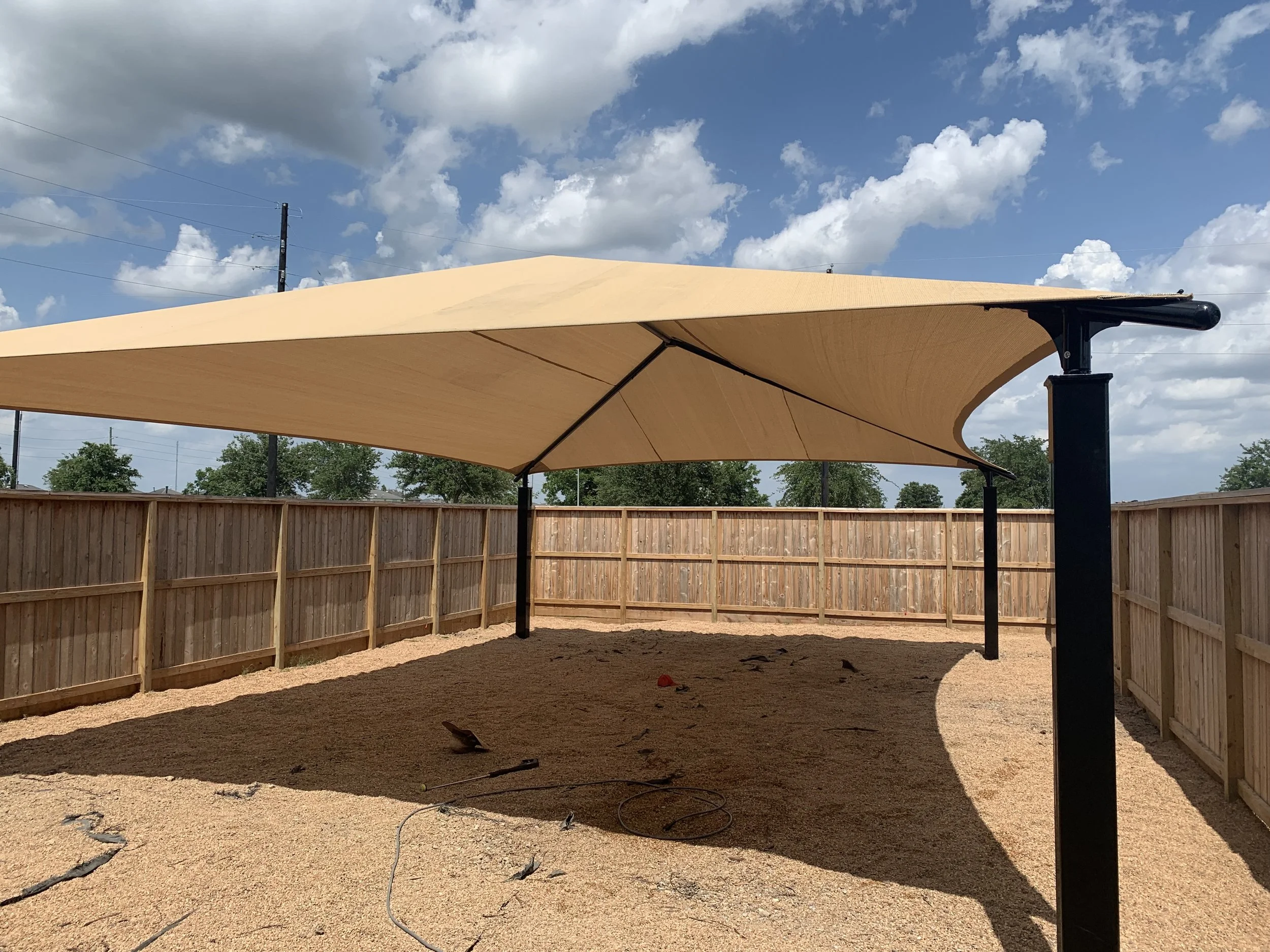 A large beige fabric canopy shade sail installed over a fenced backyard with sandy ground, some tools, and tree shadows, under a partly cloudy sky.