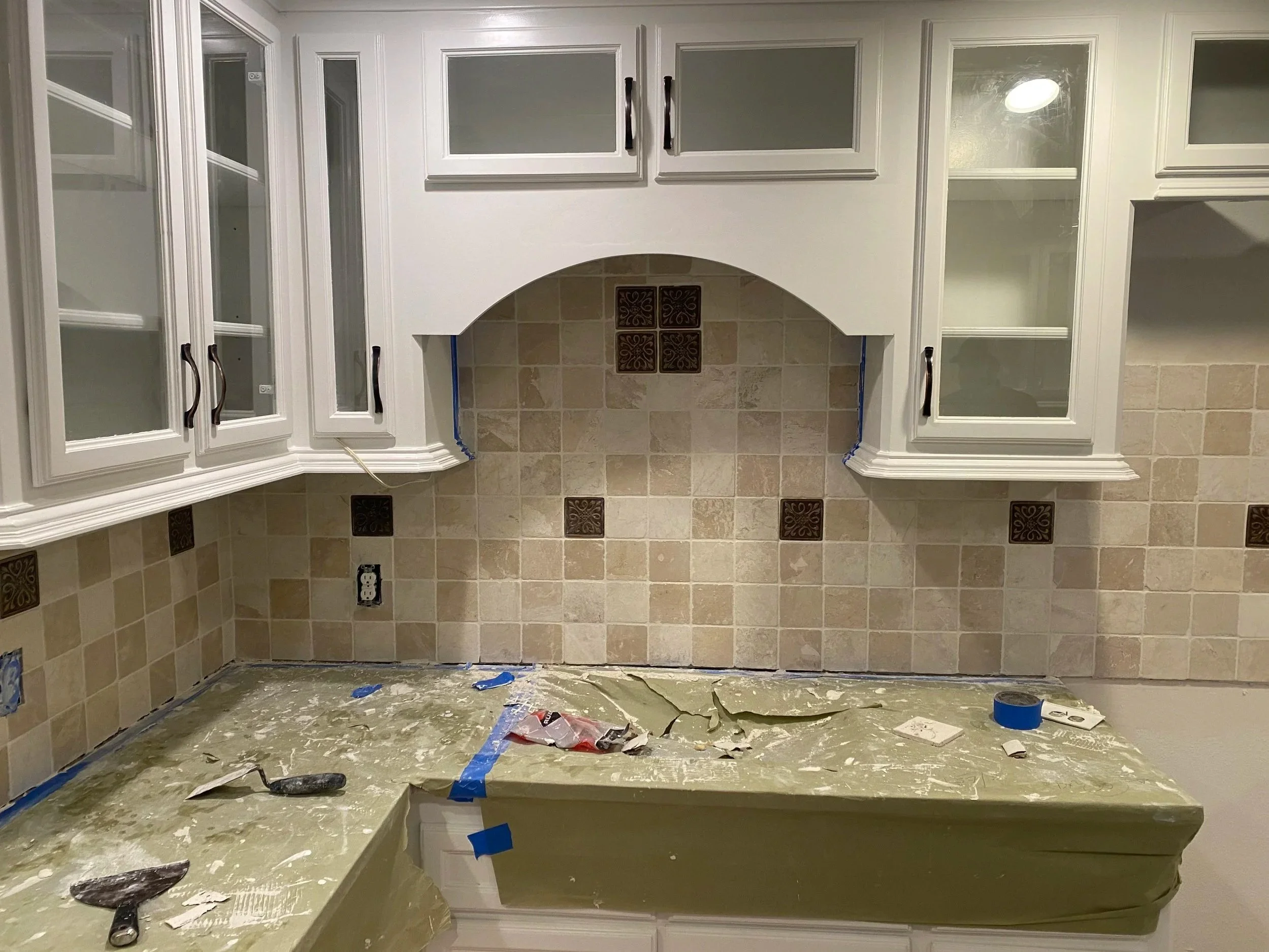 Kitchen wall with cream-colored tile backsplash, white cabinets, and a marble countertop under construction. Tools and blue painter's tape are on the counter.