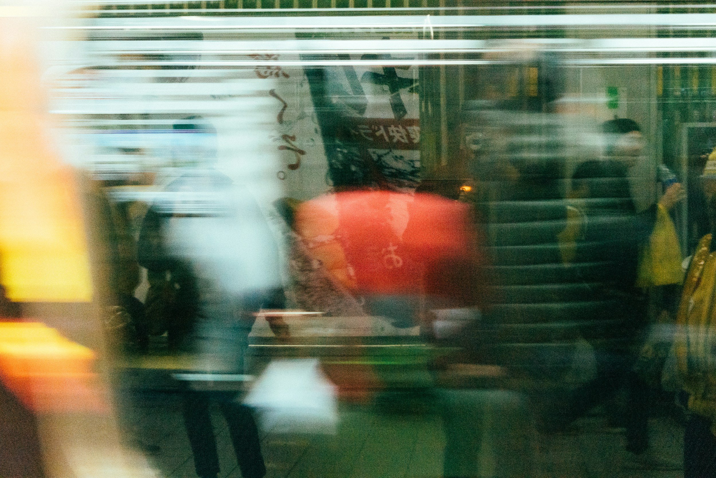 Blurred view of a city street seen through a shop window, with reflections of people passing by and storefront signs.