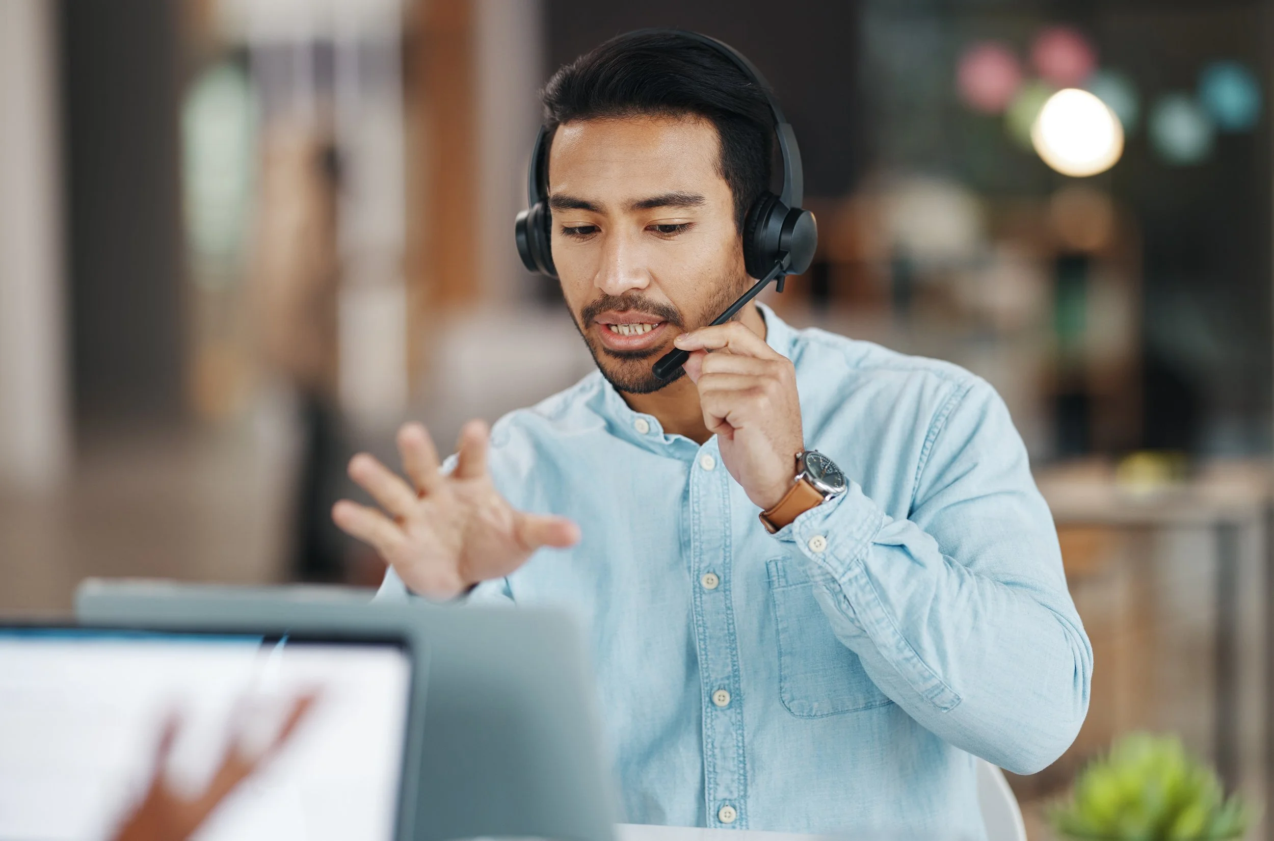 A man wearing a light blue shirt and a wristwatch, using a black headset with a microphone, appears to be speaking or explaining something while looking at a laptop in an indoor setting.