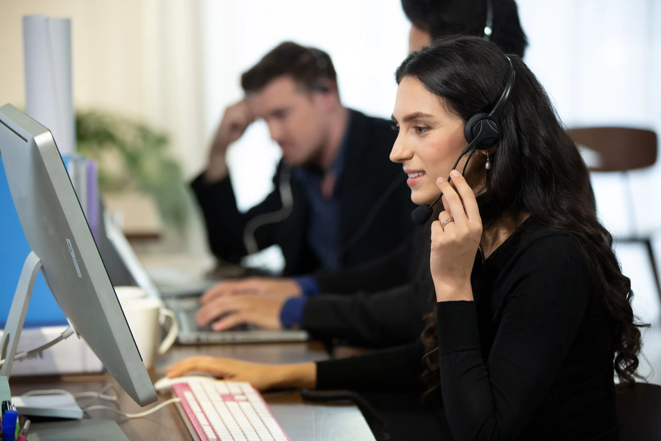 A woman with long dark hair wearing a headset, seated at a desk working on a computer with a man in the background, also working on a computer, in an office setting.