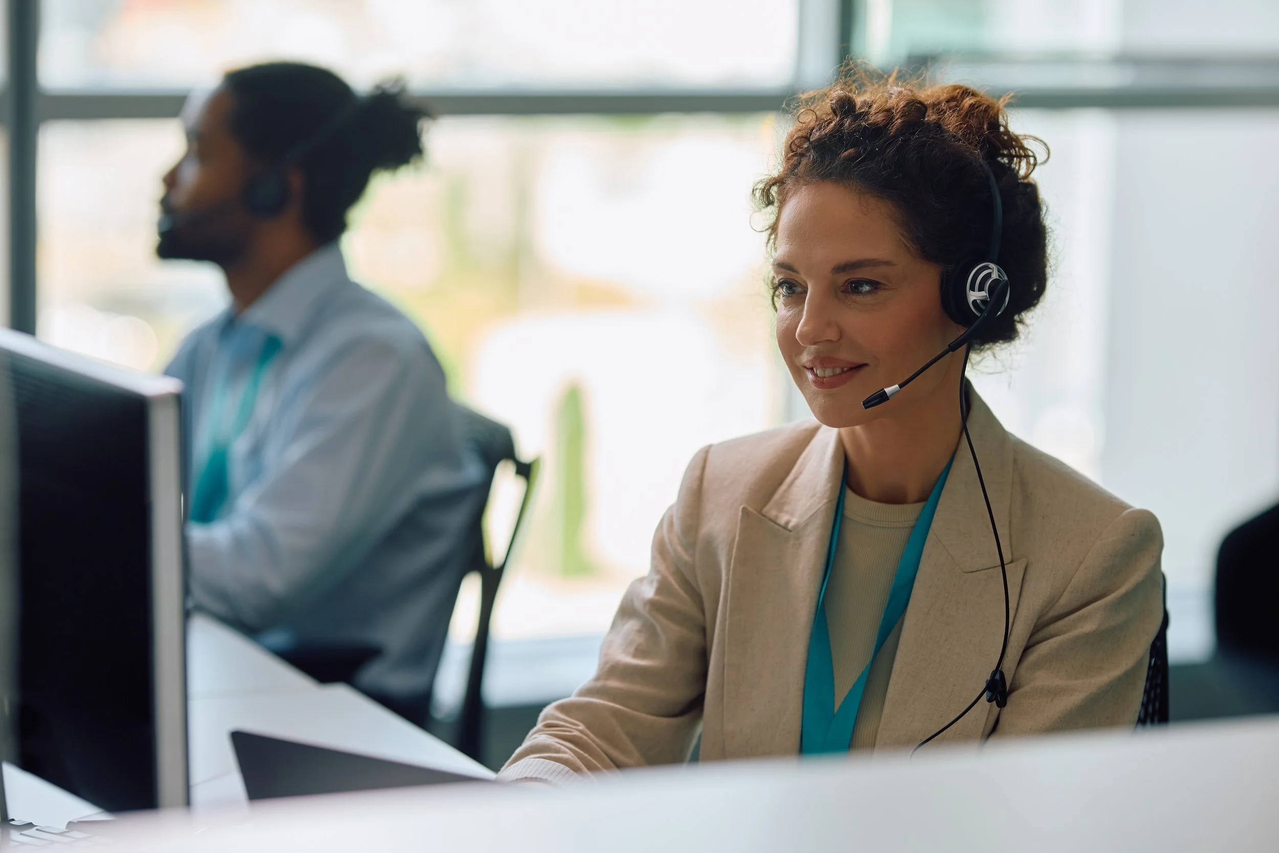 A woman wearing a beige blazer with a headset working at a computer in an office.