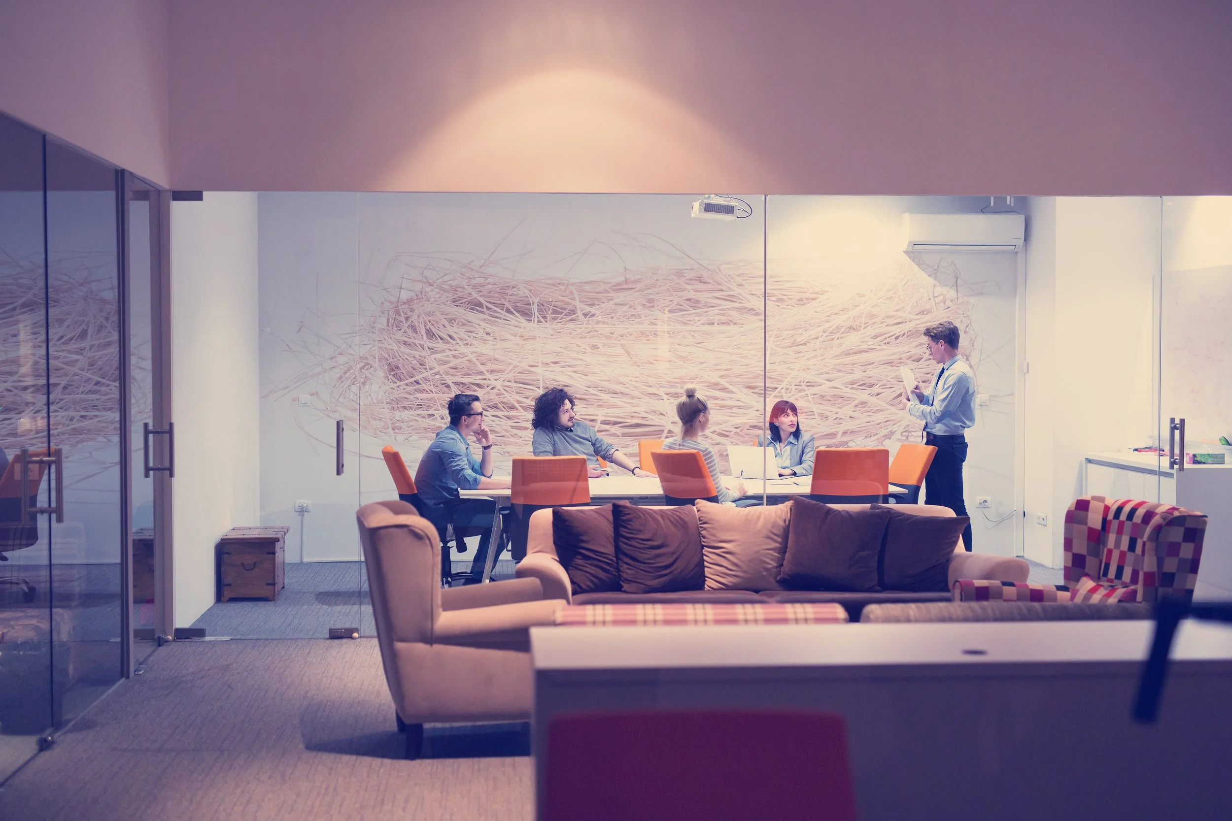 A group of five people sitting around a conference table in a modern office meeting room, with a man standing and reading from a paper. The room has a large wall mural with a pink, abstract design.