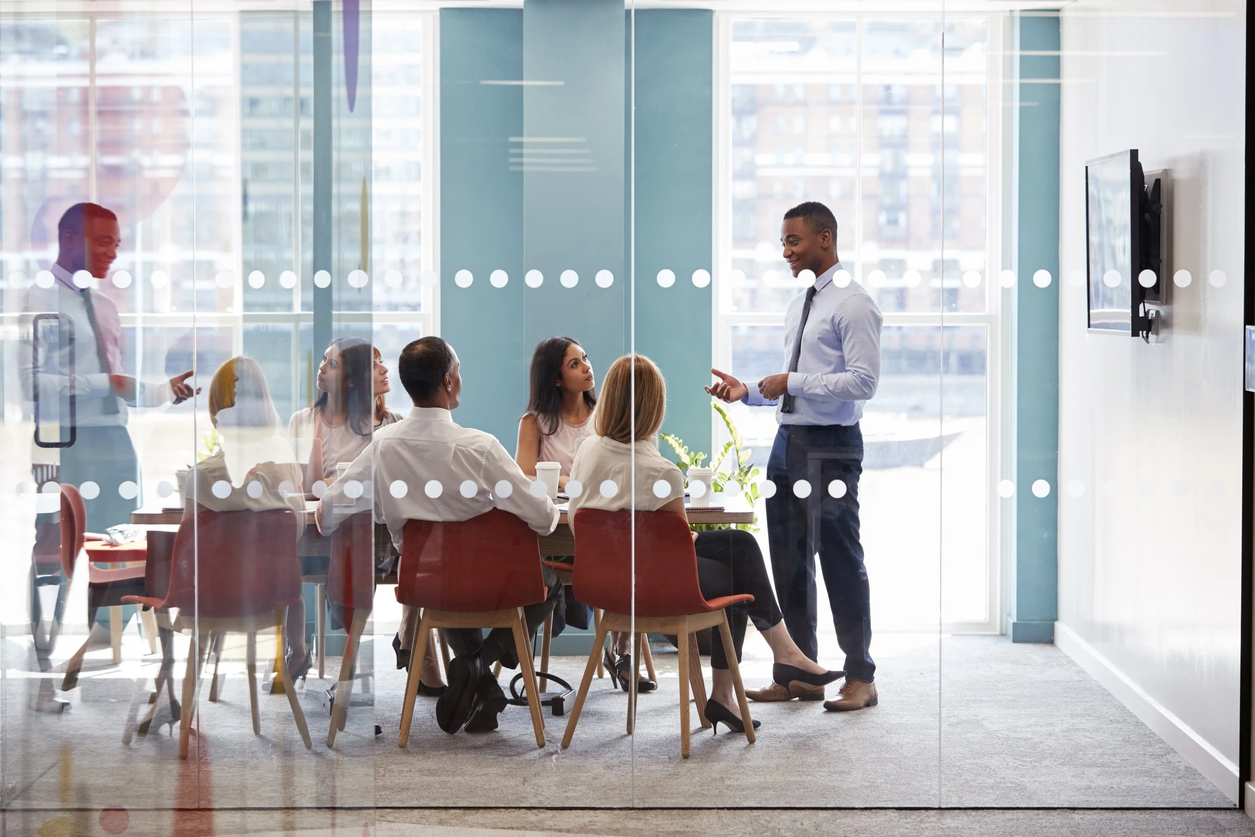Business meeting in a modern glass conference room with six diverse professionals listening to a speaker in formal business attire.