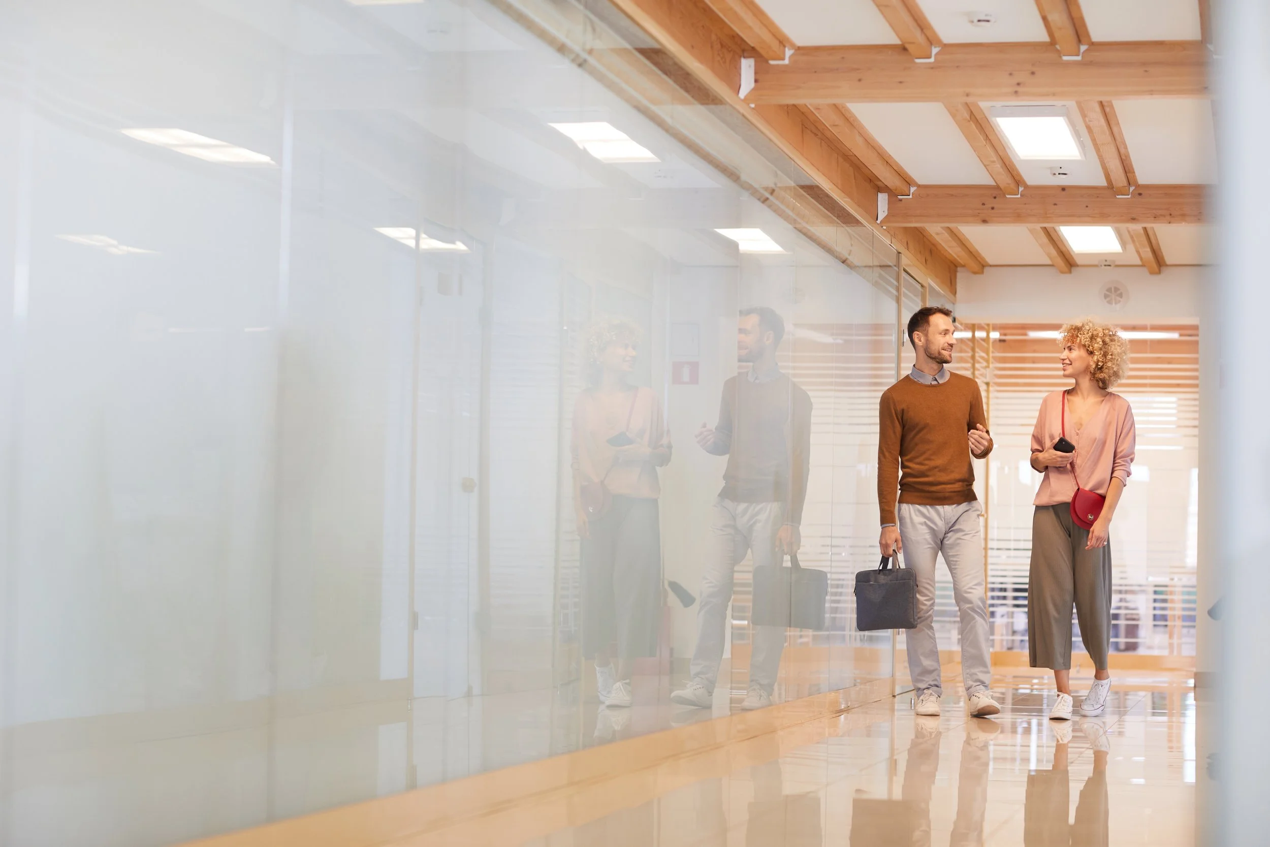 Four professionals walking and conversing in a modern office corridor, reflected in glass walls, with wooden beams and ceiling lights.