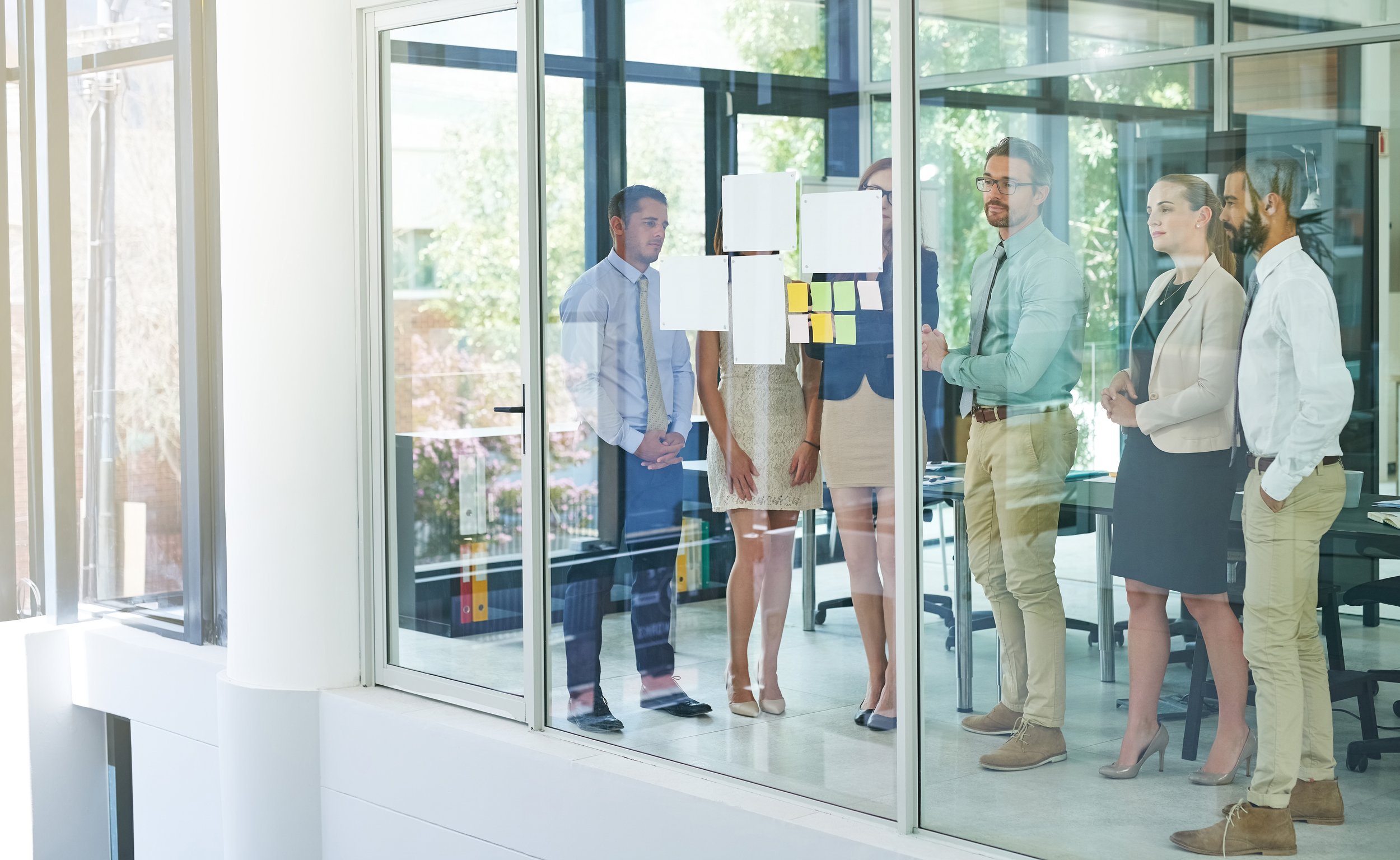 Group of six professionals standing and discussing in a glass-walled conference room, with sticky notes on the glass and cityscape visible outside.