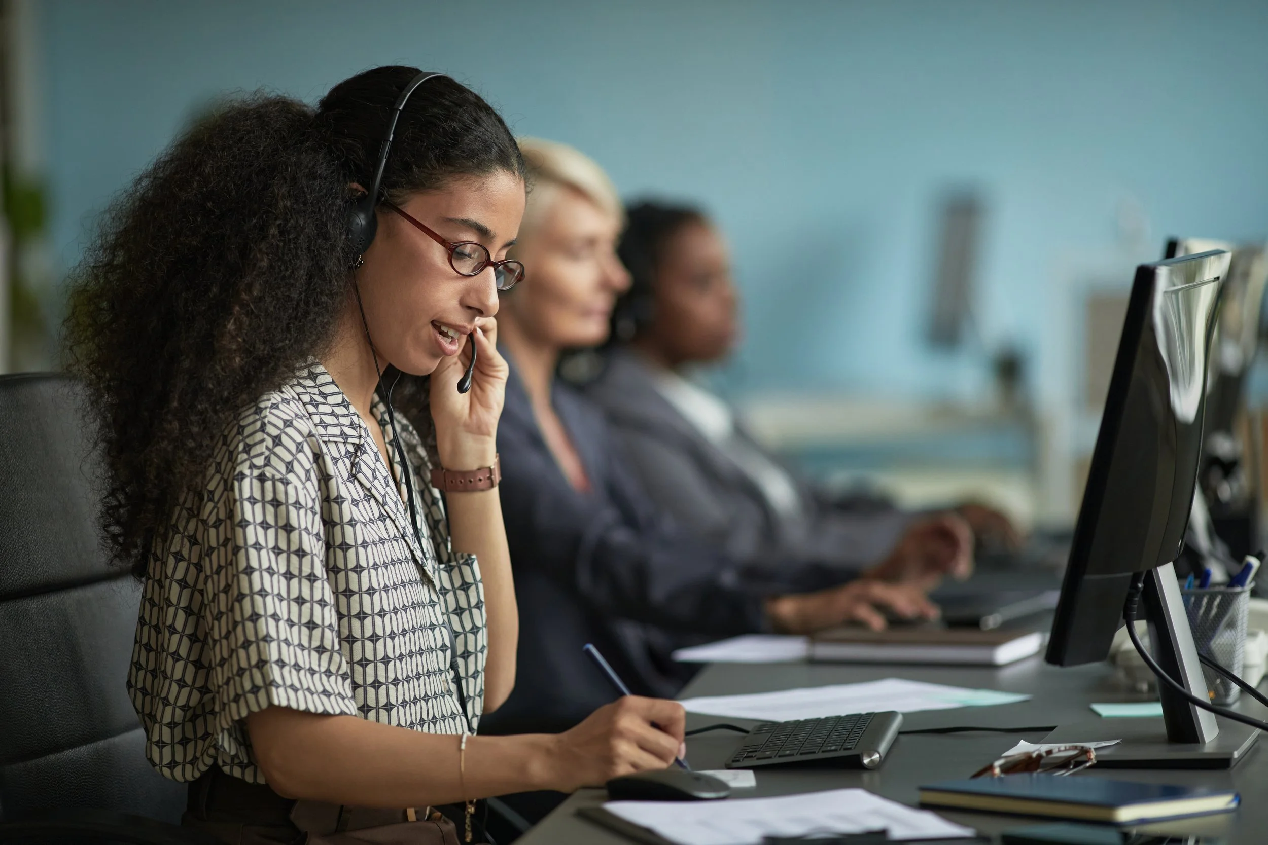 Three women working at desks in an office, with the woman in the foreground wearing headphones and talking on the phone.
