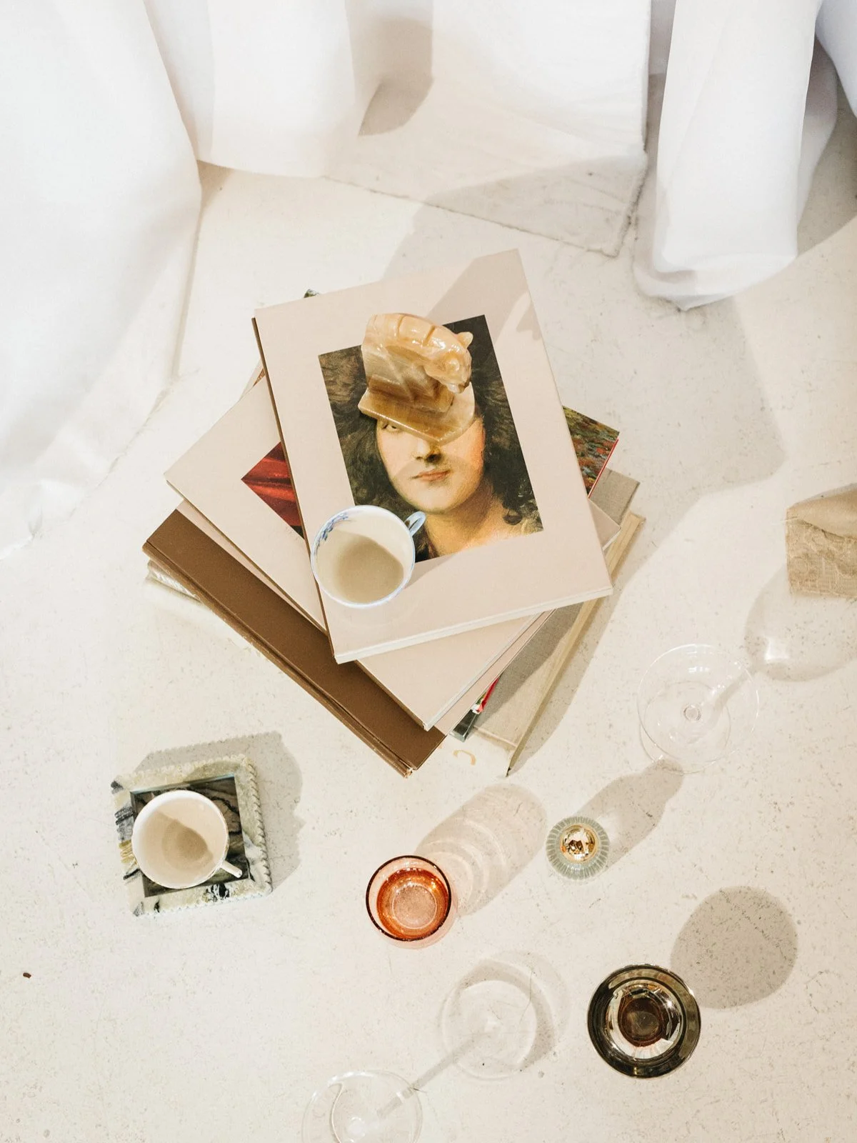 A top-down view of a table with stacked art and coffee books, a piece of carved stone sculpture, a cup of coffee, and several wine glasses and candles on a white surface.