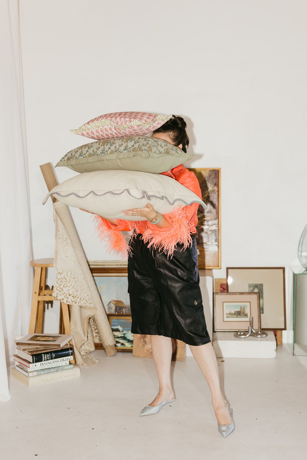 A woman is holding three decorative pillows in front of her face, standing in an art studio or gallery with framed artwork and books in the background.