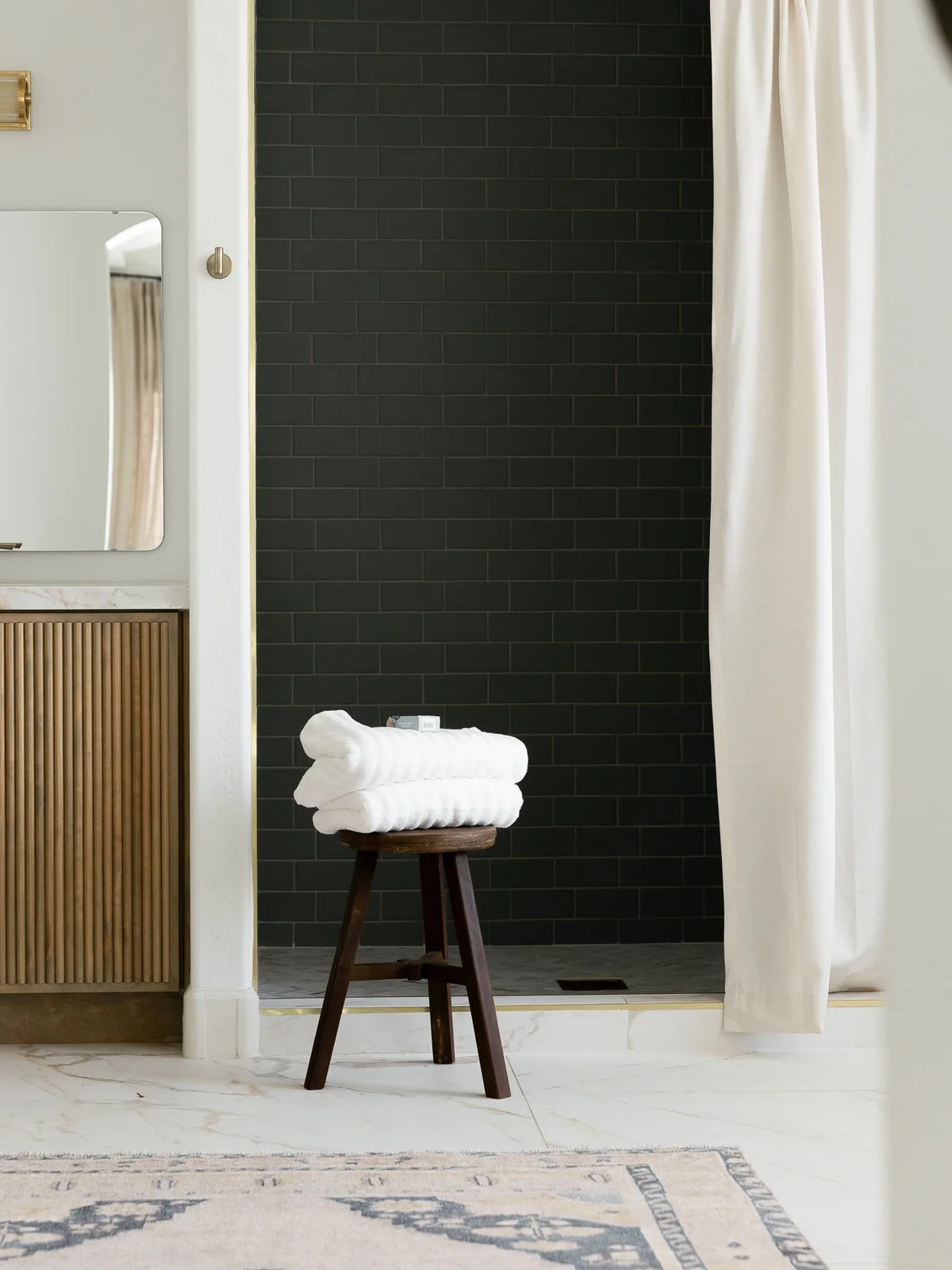 A bathroom with a black-tiled shower area, a white curtain, a wooden stool with folded white towels on top, a marble countertop, a mirror, and beige and white decor.