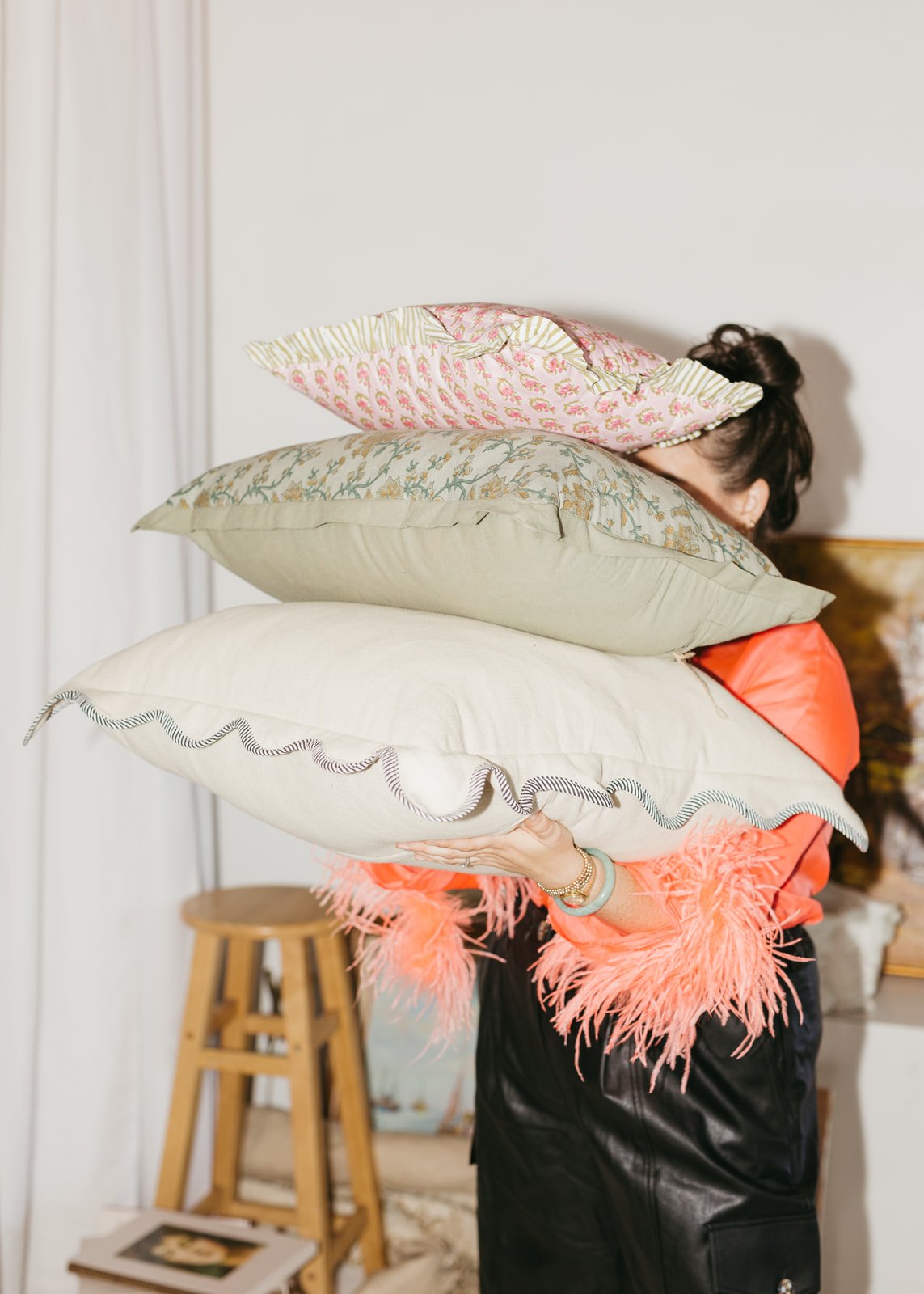 Person holding a stack of decorative pillows in a room with a wooden stool and a small table.