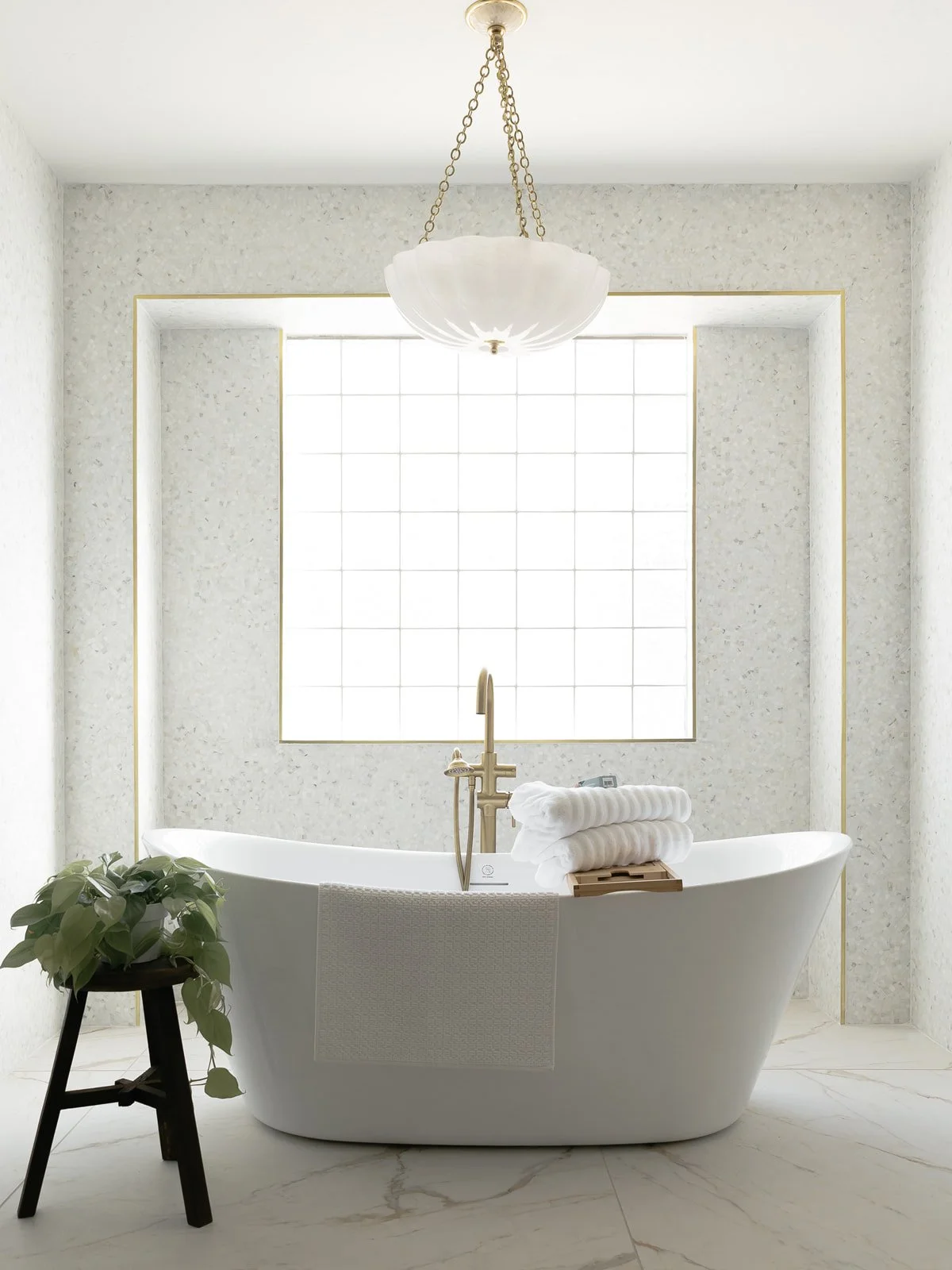 A modern white bathtub with a gold faucet in a bright bathroom with large window, hanging white chandelier, a potted plant, and towels.