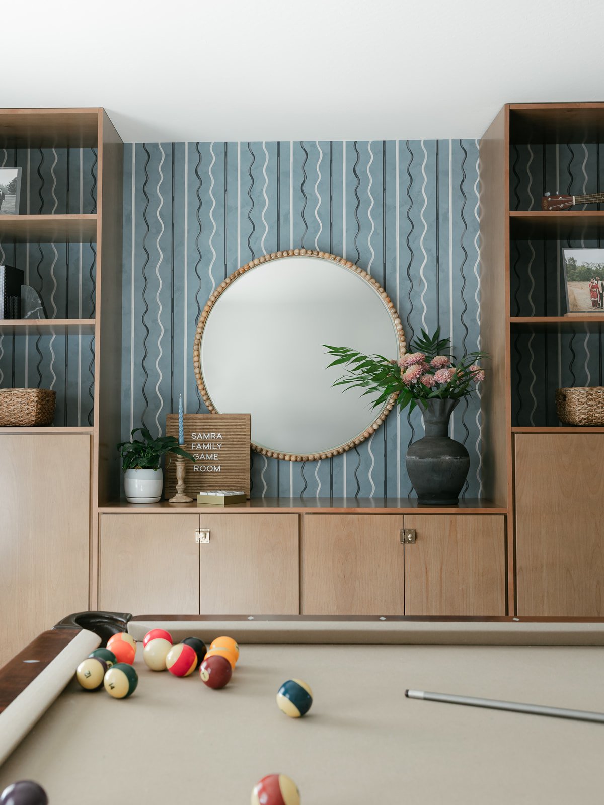 A room decorated with a large round mirror, a floral arrangement in a dark vase, a potted plant, and a sign that reads 'SAMRA FAMILY GAME ROOM.' A pool table with some pool balls and cue stick in the foreground.
