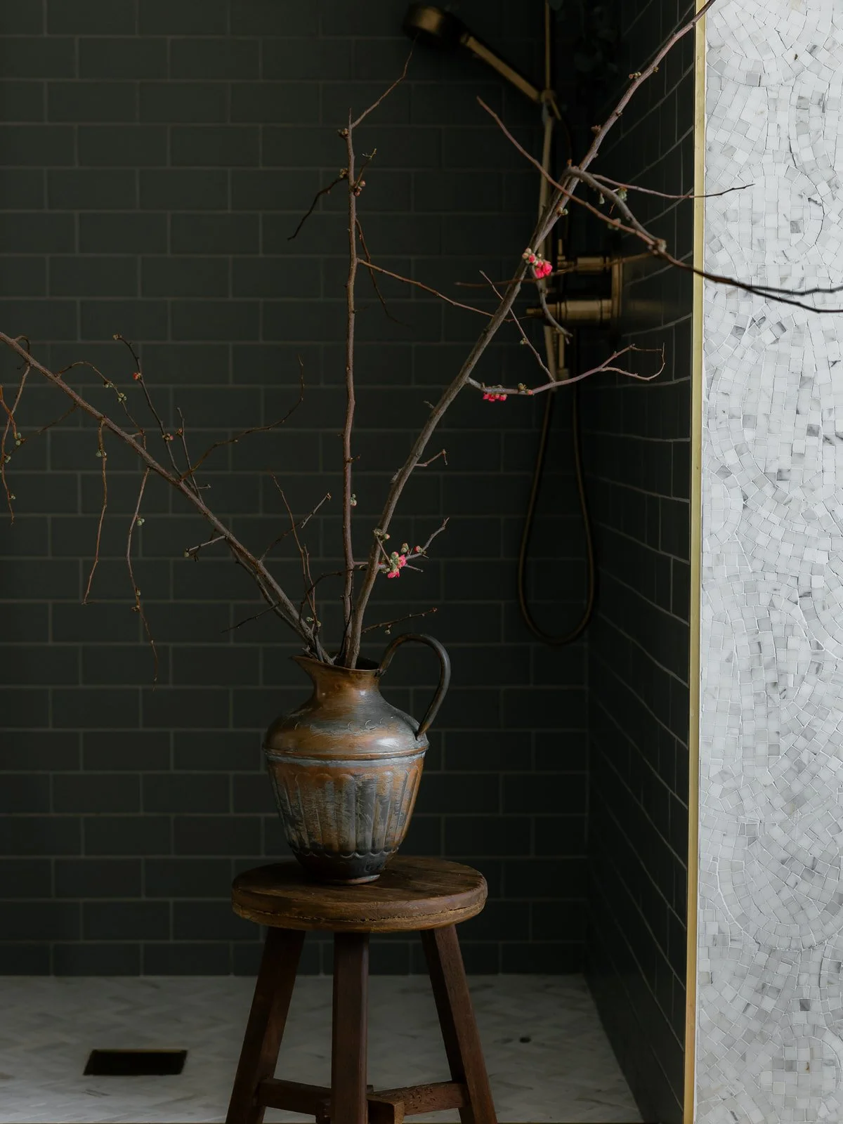 A brown ceramic vase with a handle, containing bare tree branches with small pink blossoms, placed on a round wooden stool against a dark tiled wall and a white mosaic wall.