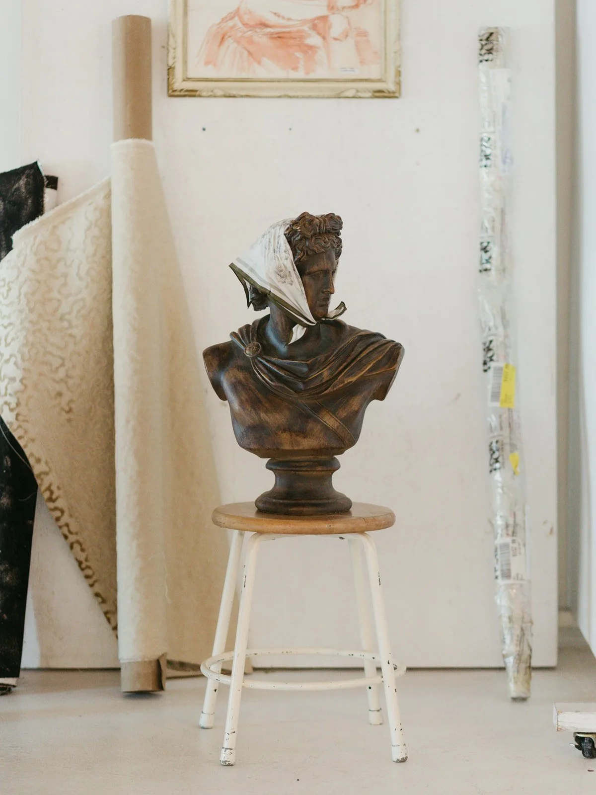 A bronze bust sculpture of a woman with a white scarf around her head, placed on a wooden pedestal inside a room with some art supplies and artwork visible in the background.
