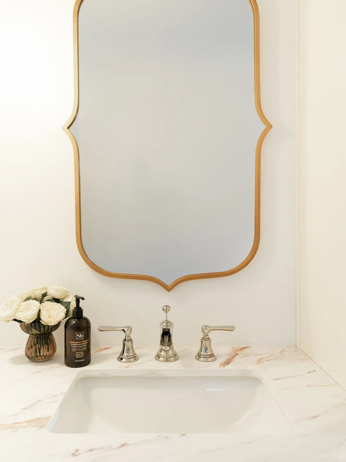 Bathroom sink with a marble countertop, silver faucet, a vase with white roses, a bottle of moisturizing lotion, and a decorative mirror above.