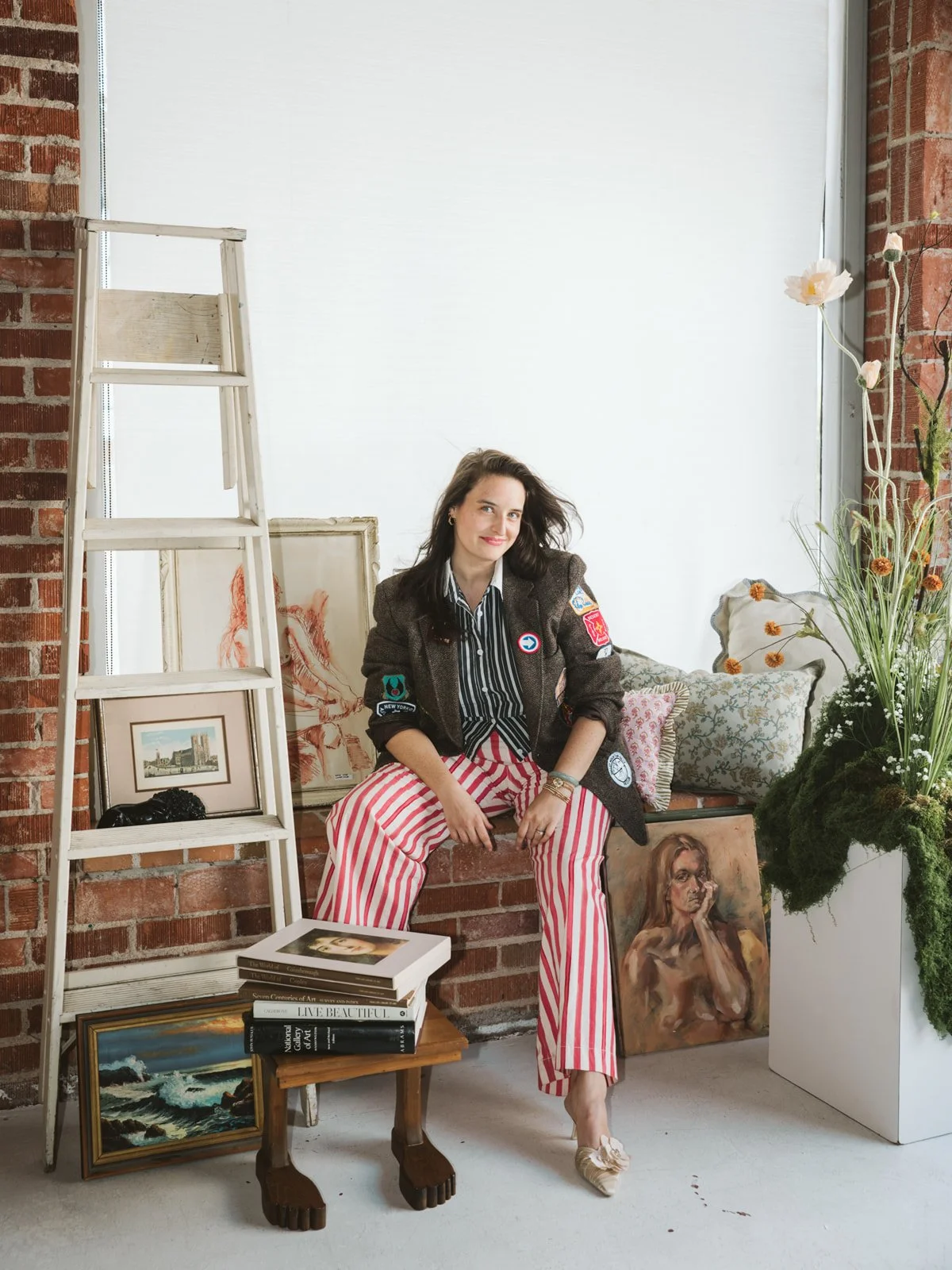A woman sits on a brick ledge surrounded by paintings, books, and plants in an art studio or gallery space.